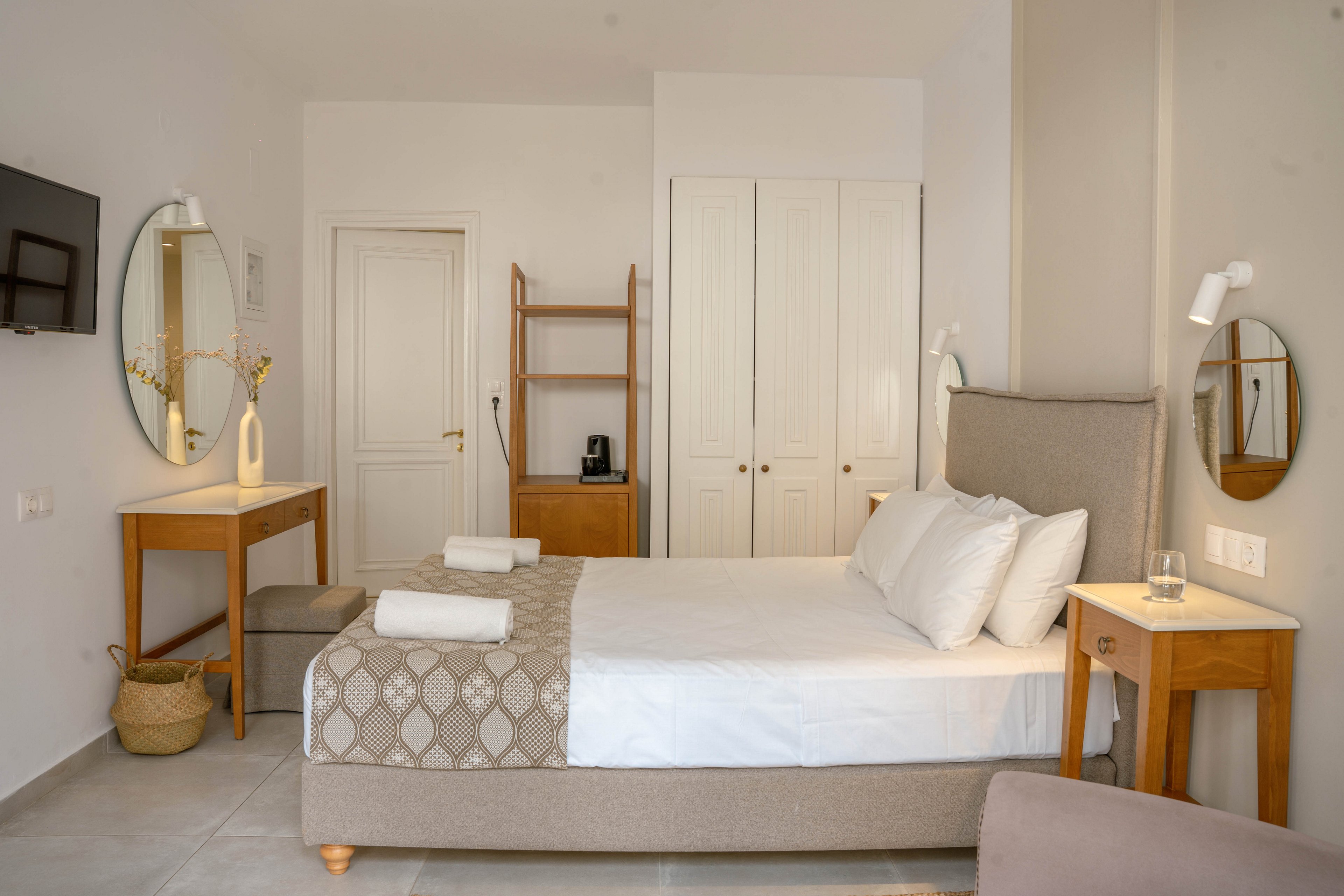 A neatly made double bed with white linens and a patterned beige throw is the focal point of this cozy bedroom. The room features light neutral walls, tiled flooring, and simple wooden furniture including bedside tables and a writing desk. A wall-mounted TV, round mirrors, and a small open wardrobe with a coffee maker add functional touches. Soft lighting and minimal decorative accents create a calm, contemporary atmosphere.