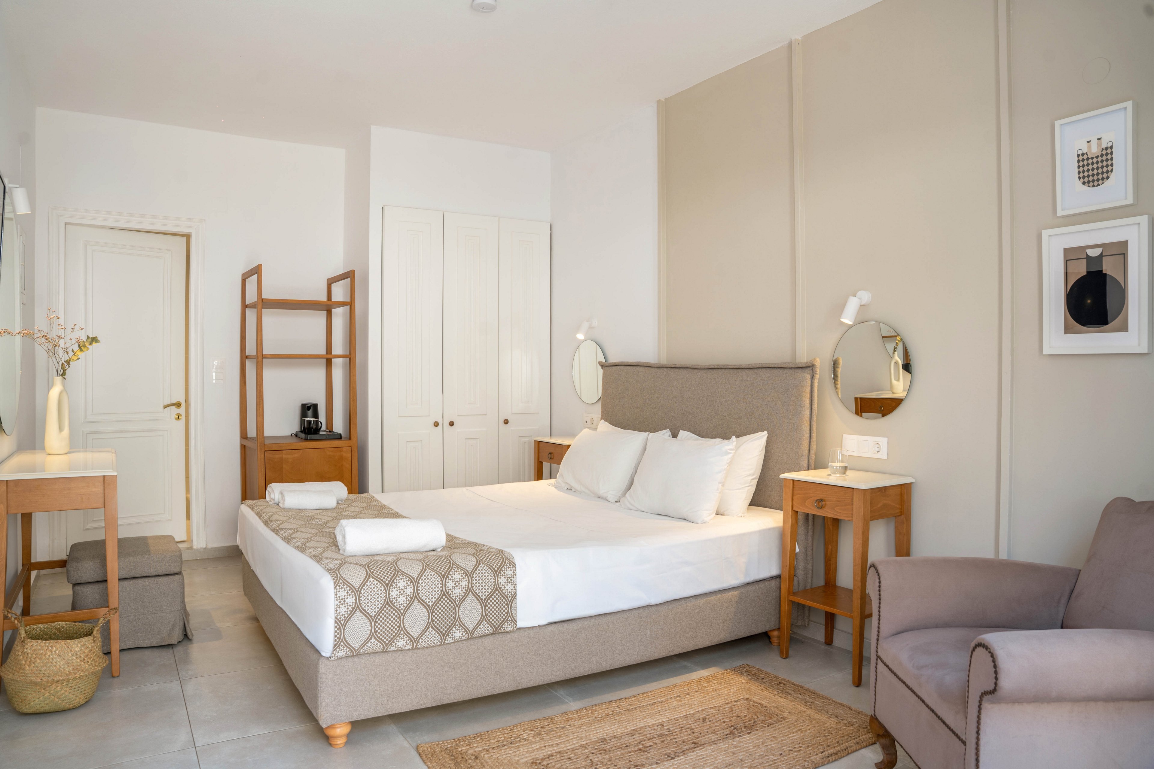 A neatly made double bed with white linens and folded towels is the focal point of this bright bedroom. The room features light neutral walls, a wooden nightstand, and a simple wooden shelving unit near a white door. A soft armchair, woven rug, and small desk with mirror add comfort and functionality. Framed abstract artwork and subtle decorative accents create a calm, minimalist aesthetic.