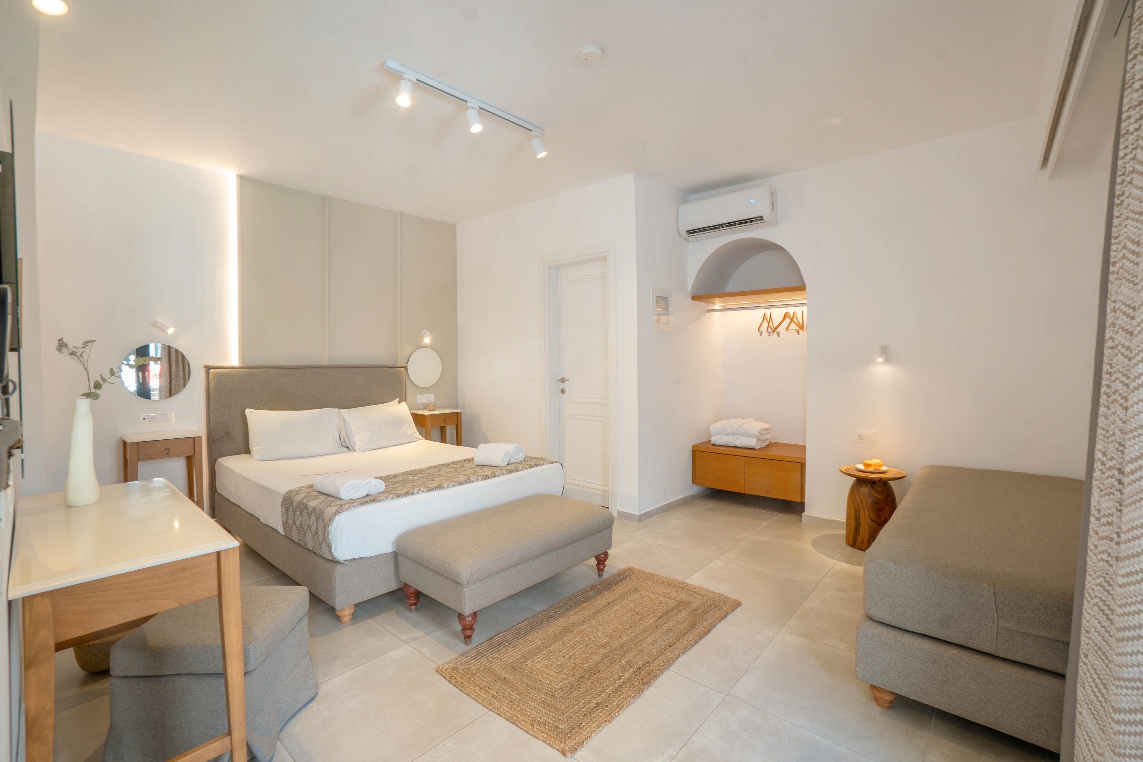 A spacious, modern hotel bedroom features a large bed with a padded headboard and matching bench. The room is decorated in neutral tones with light wood furniture, including a desk, chair, and side tables. Soft lighting, a wall-mounted air conditioner, and an open wardrobe area contribute to a clean, minimalist aesthetic. A woven rug and simple accessories add warmth and comfort to the contemporary space.