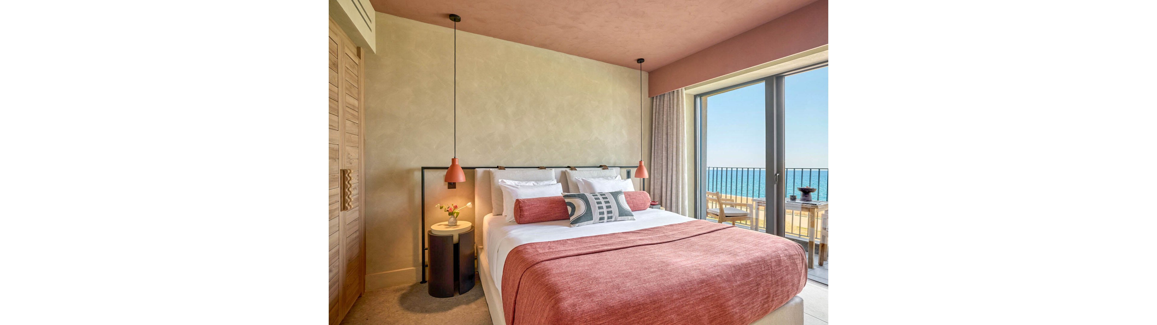 Stylish hotel bedroom features a neatly made double bed with pink and white linens and decorative cushions. The room includes modern pendant lights, a small round side table with flowers, and soft neutral walls. Large floor-to-ceiling windows open onto a balcony with outdoor seating and a clear view of the sea and blue sky. The overall design feels contemporary, cozy, and inviting for a relaxing seaside stay.