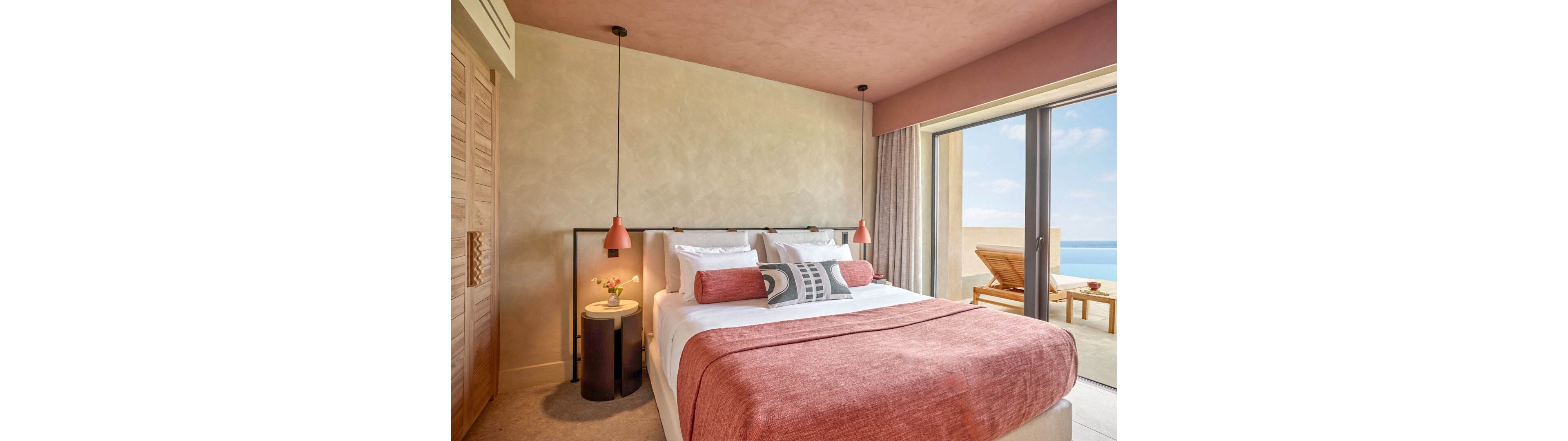 A contemporary bedroom features a neatly made double bed with layered pillows and a soft pink throw. Pendant lights hang on either side above small bedside tables, one holding a vase with flowers. Large sliding glass doors open to a terrace with a sun lounger and table overlooking a bright blue sea and sky. Warm neutral walls and blush tones create a calm, inviting atmosphere.