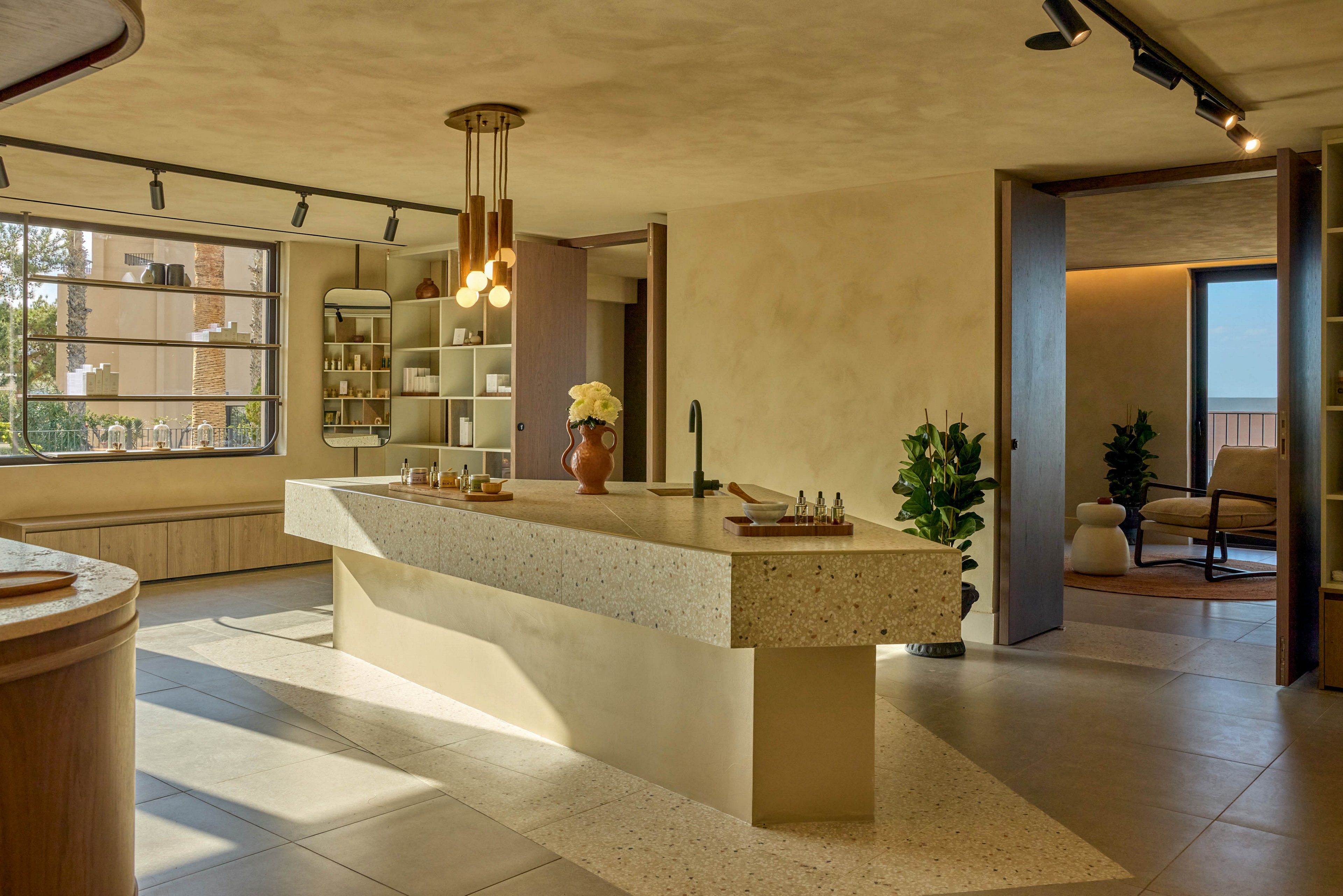 A contemporary kitchen features a large terrazzo island as its central focal point. Warm natural light streams through a wide window, illuminating minimalist cabinetry and open shelving with neatly arranged objects. Pendant lights hang above the island, while track lighting runs along the ceiling for additional illumination. An adjacent sitting area and potted plants add a cozy, residential feel to the modern interior.