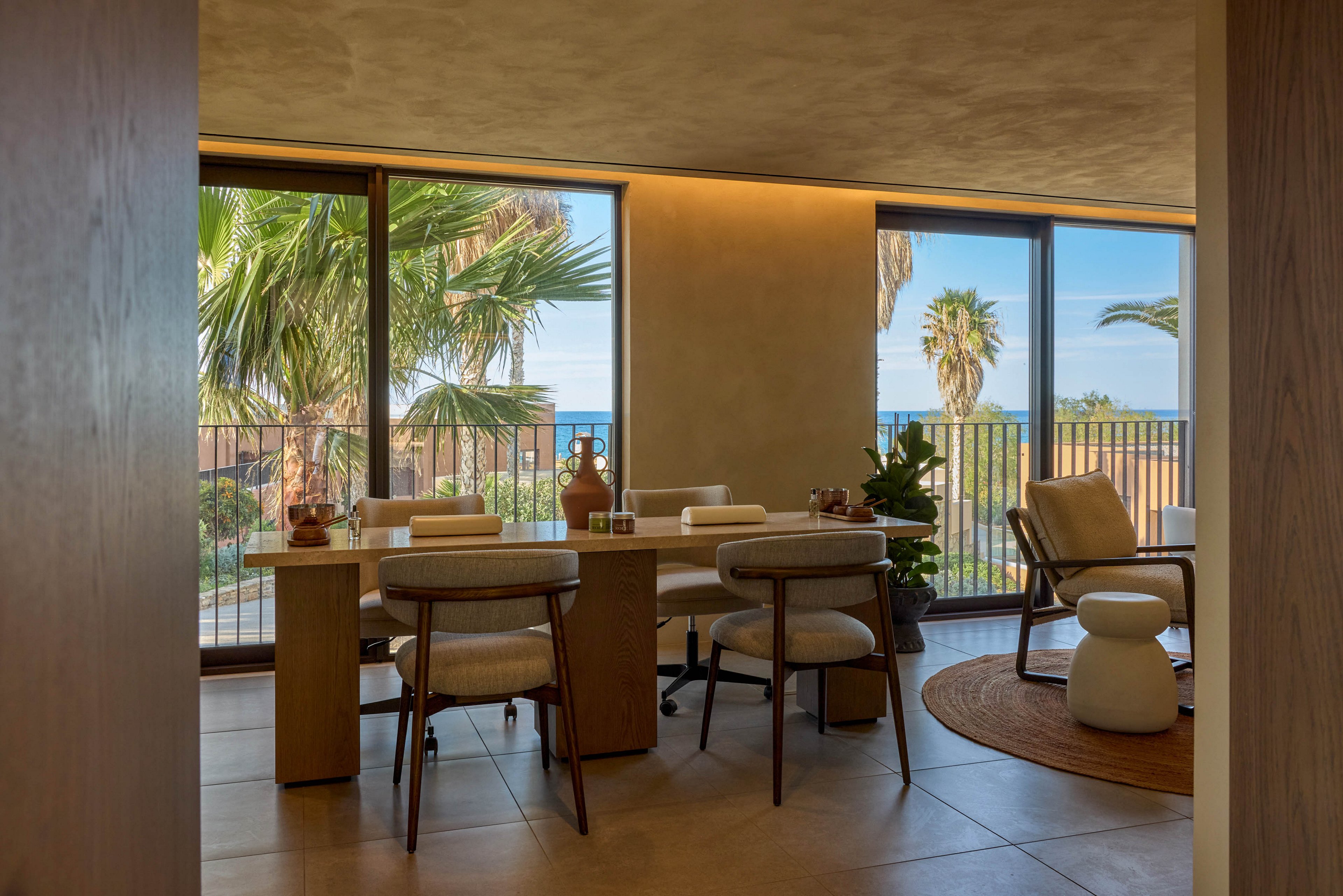A contemporary dining area features a wooden table surrounded by cushioned chairs. Large floor-to-ceiling windows open to a balcony with lush palm trees and a distant ocean view. Warm, indirect lighting and neutral-toned walls create a cozy, sophisticated atmosphere. Minimalist decor, indoor plants, and a lounge chair on a round rug enhance the relaxed coastal style.