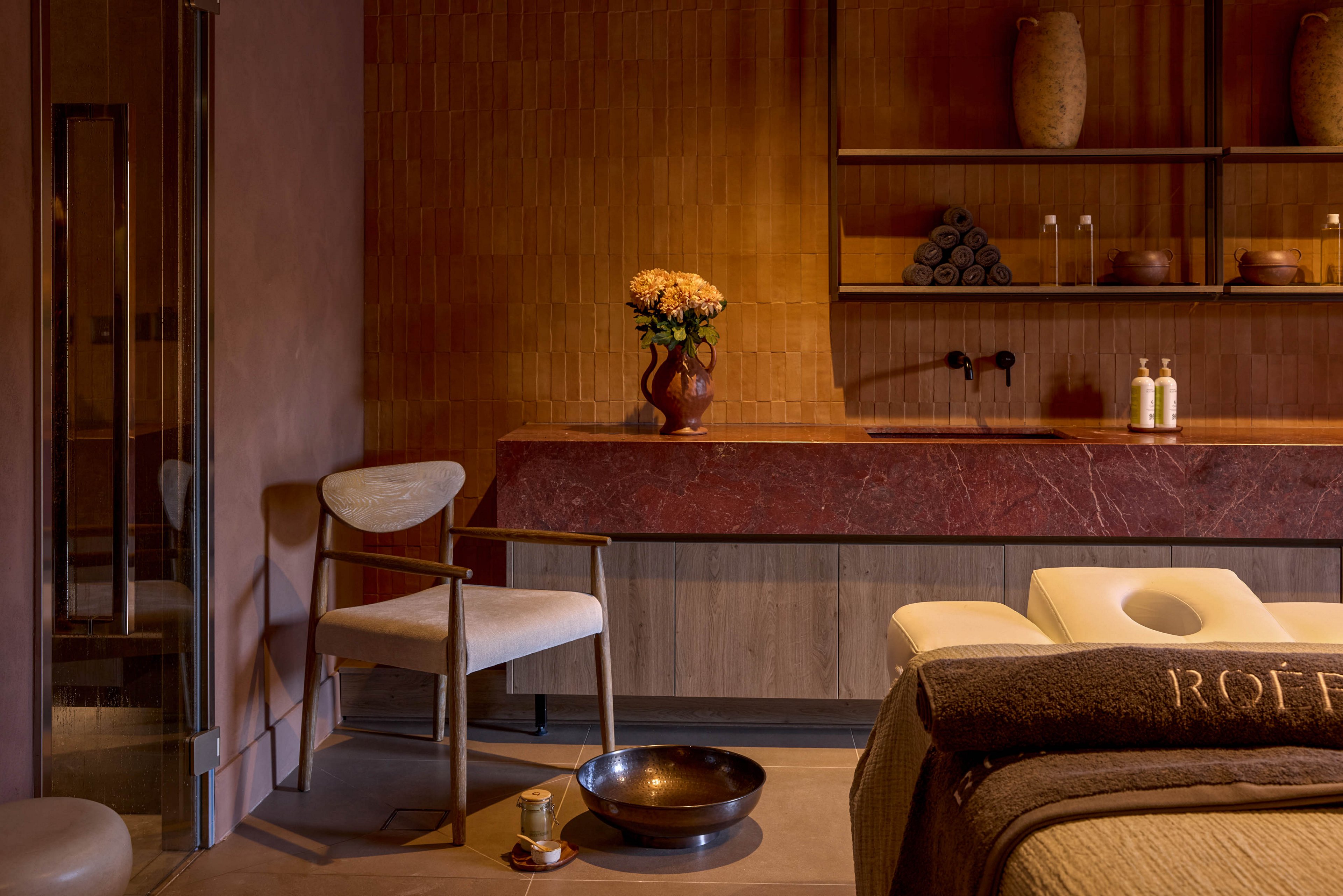 A cozy spa treatment room features a massage table and a modern armchair under warm ambient lighting. The space includes a long stone counter with decorative vases, folded towels, and two pump bottles neatly arranged. A metal bowl and small container sit on the floor, suggesting a foot bath or ritual treatment. Earthy tones of brown, beige, and terracotta create a calm, luxurious atmosphere.