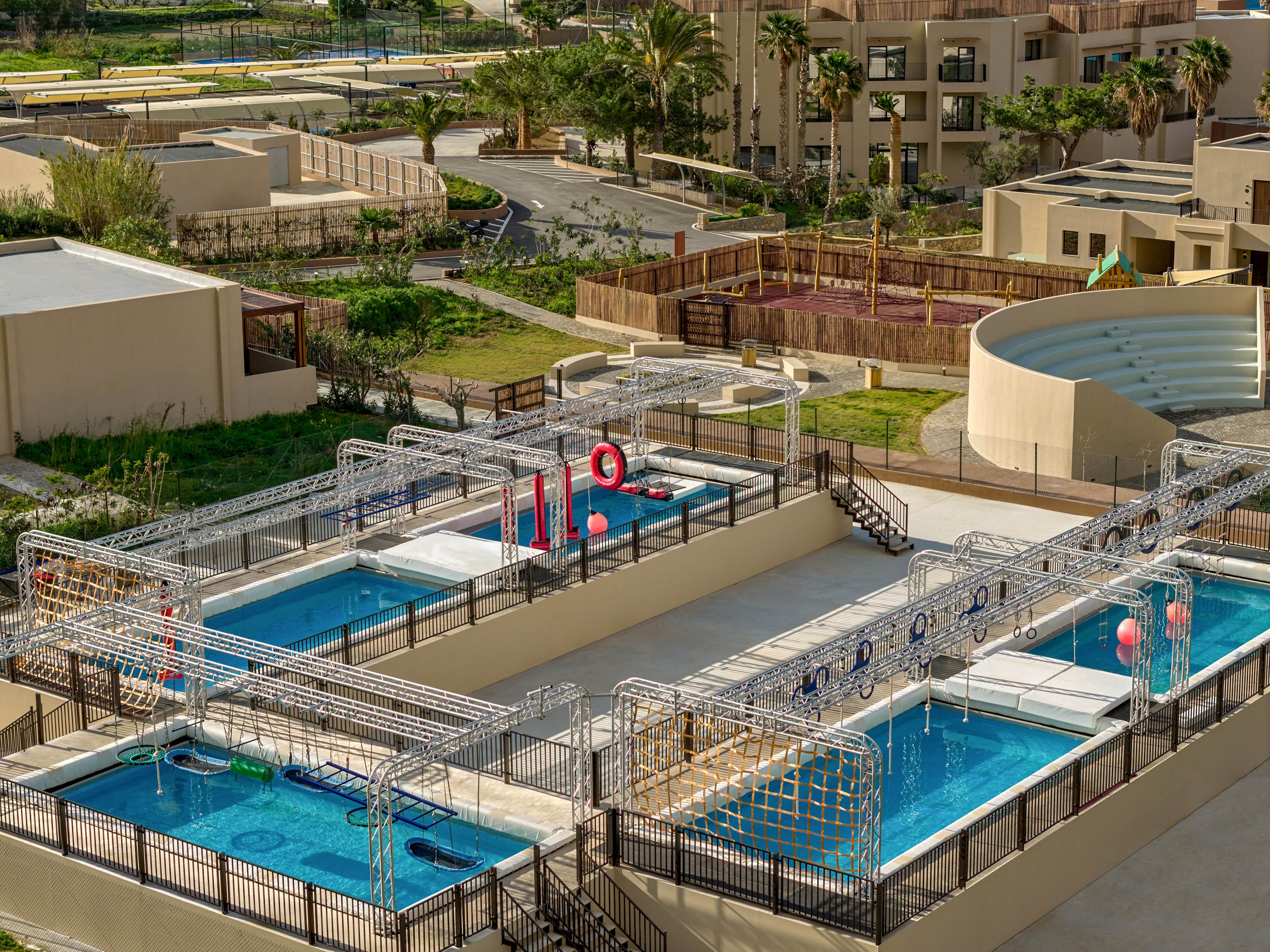 An elevated view shows multiple rectangular outdoor swimming pools at a resort or recreation complex. Metal obstacle course frames span the pools, with hanging rings, ropes, and floating platforms for water activities. Surrounding the pools are beige buildings, landscaped greenery, and winding pathways. The scene is brightly lit, suggesting a warm, sunny day suitable for leisure and play.