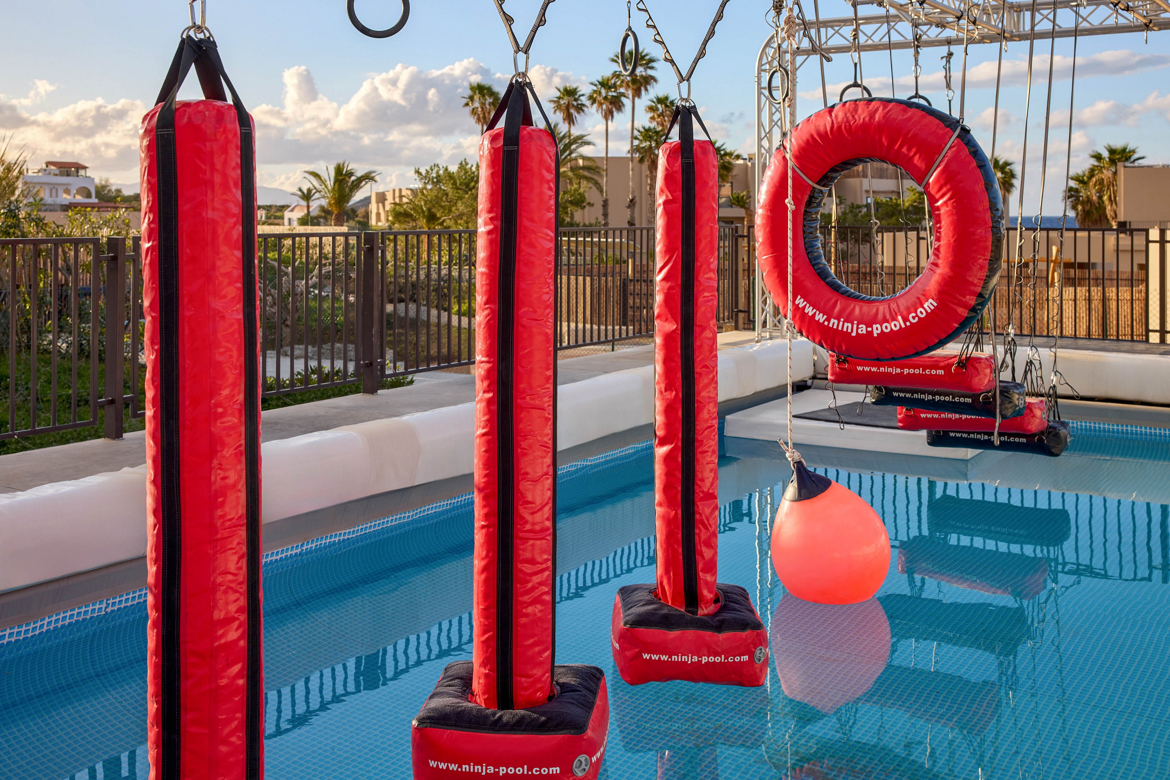An outdoor inflatable obstacle course is set up over a bright blue pool. Tall red padded pillars and a large circular ring hang from metal frames above the water. A glowing red buoy floats in the pool, adding to the playful atmosphere. The scene is surrounded by fencing, palm trees, and warm evening light, suggesting a resort or recreational park setting.