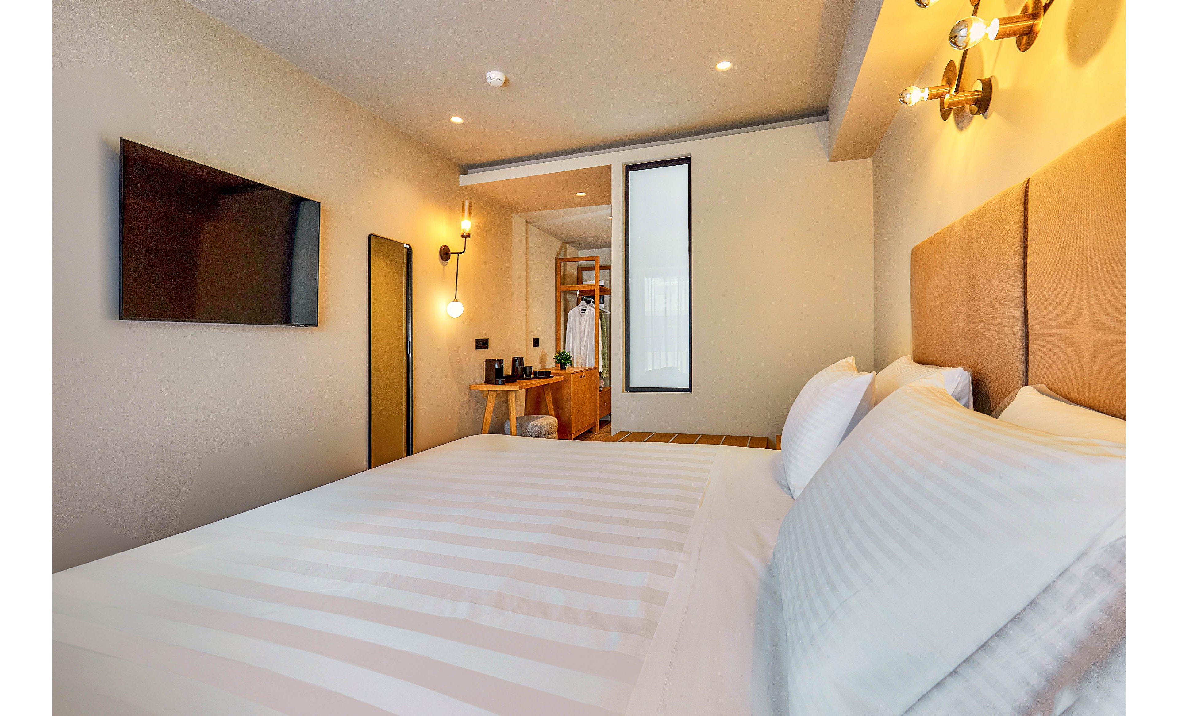 A contemporary hotel room features a large, neatly made bed with white linens and a padded headboard. The space includes a wall-mounted flat-screen television, warm lighting fixtures, and a small open wardrobe area with hangers and amenities. The room is decorated in neutral tones, creating a calm and inviting atmosphere.