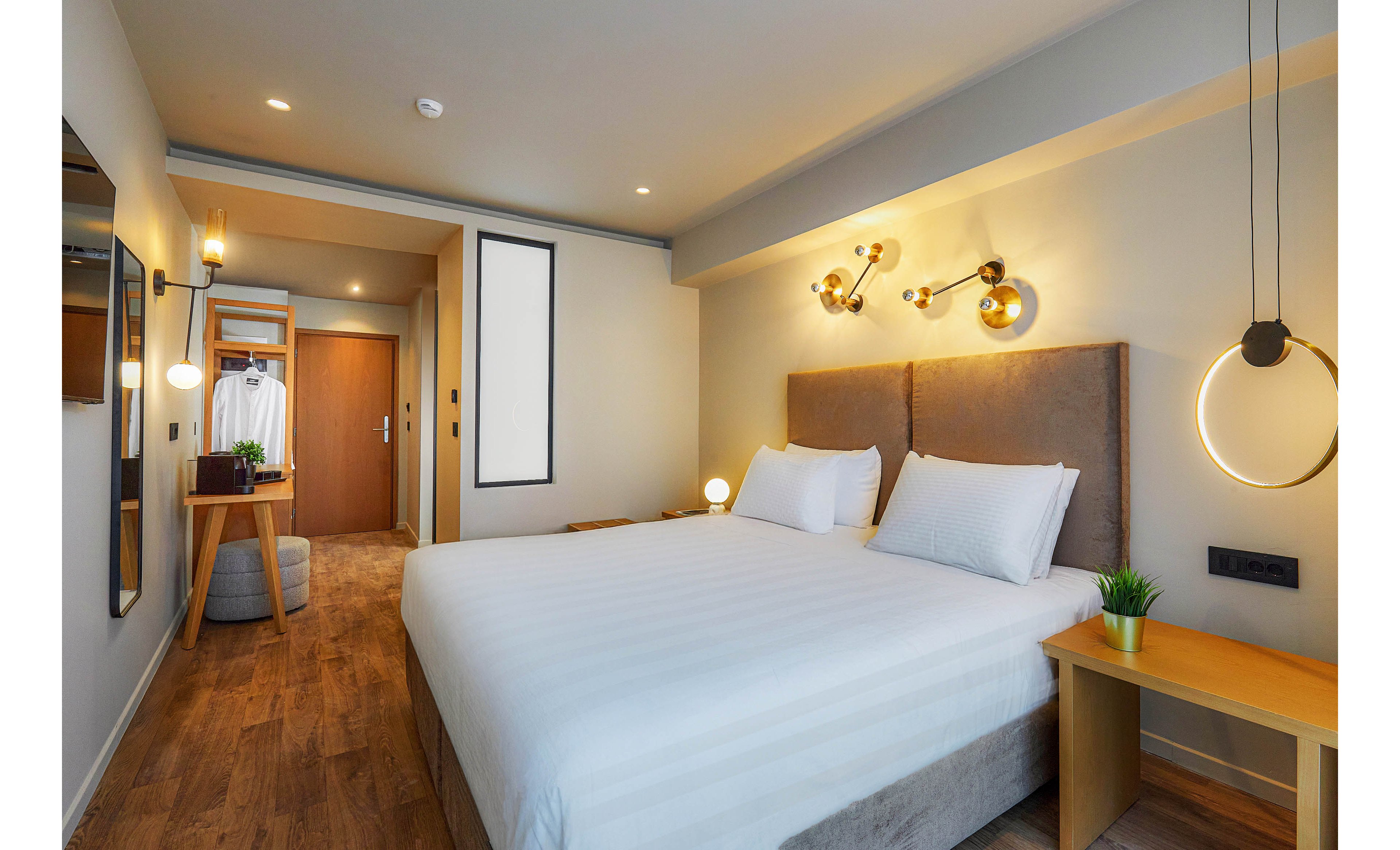 A contemporary hotel room features a large double bed with crisp white linens and plush headboard. The space is illuminated by stylish wall and pendant lights, creating a cozy and inviting atmosphere. Wooden flooring, a small desk, and a wardrobe with hanging clothes add to the functional yet elegant design. Decorative touches include a small green plant on the bedside table and minimalist wall art.