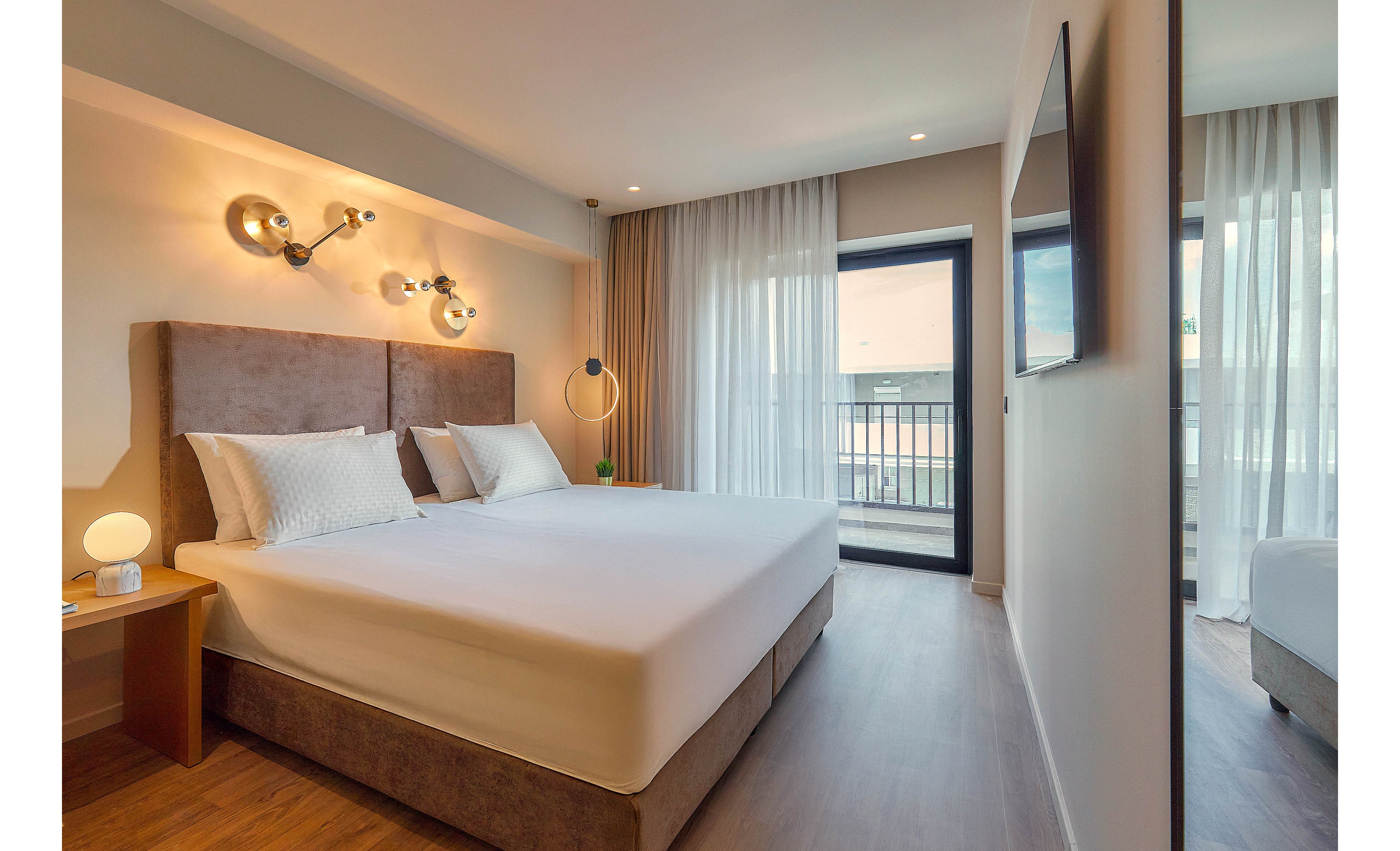 A contemporary hotel room features a spacious double bed with a padded headboard and crisp white linens. The room is illuminated by stylish wall-mounted lights and a unique floor lamp, with natural light streaming in from a balcony door and large window. Minimalist decor, neutral tones, and wood flooring create a calm and inviting atmosphere. A bedside table holds a small lamp, and a flat-screen TV is mounted on the wall.