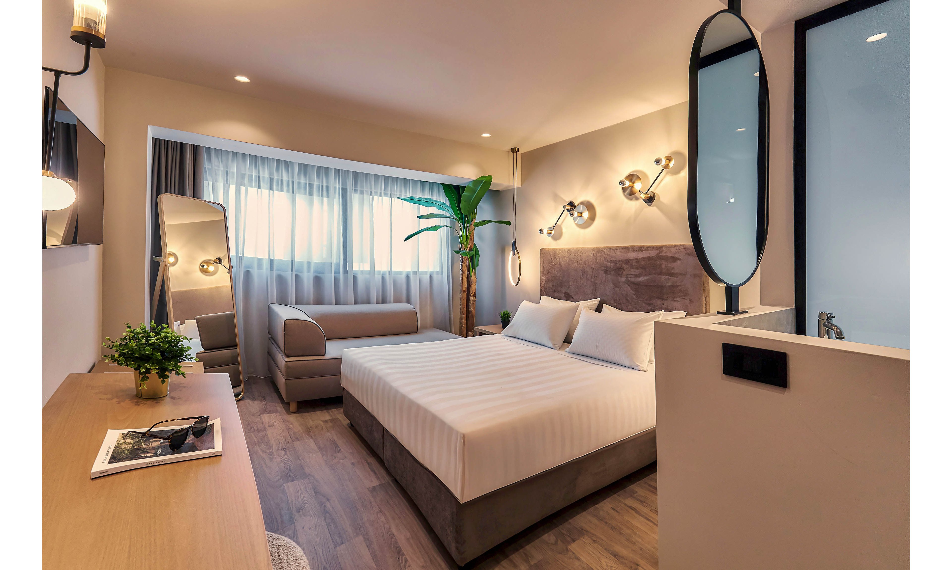 A contemporary hotel room features a large king-size bed with crisp white linens and plush pillows. The space is illuminated by stylish wall-mounted lights and natural daylight from a wide window with sheer curtains. A sleek desk, decorative plant, and a comfortable sofa add to the inviting atmosphere, while a tall mirror and minimalist decor enhance the modern aesthetic.