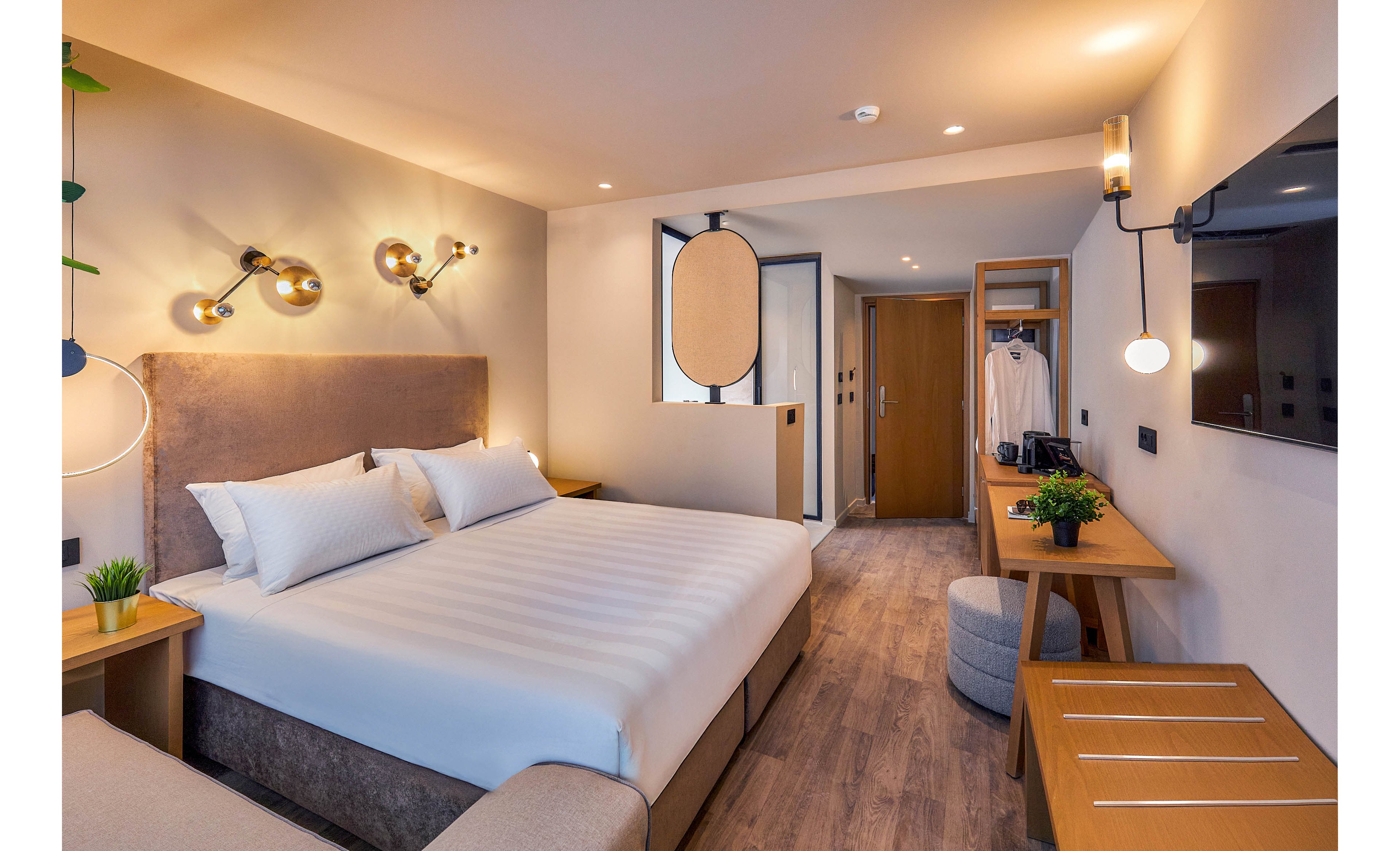 A contemporary hotel room features a large king-size bed with crisp white linens and plush pillows. The space includes a wooden desk, wall-mounted TV, and open wardrobe with hanging clothes. Warm lighting, minimalist decor, and neutral tones create a welcoming and comfortable atmosphere.