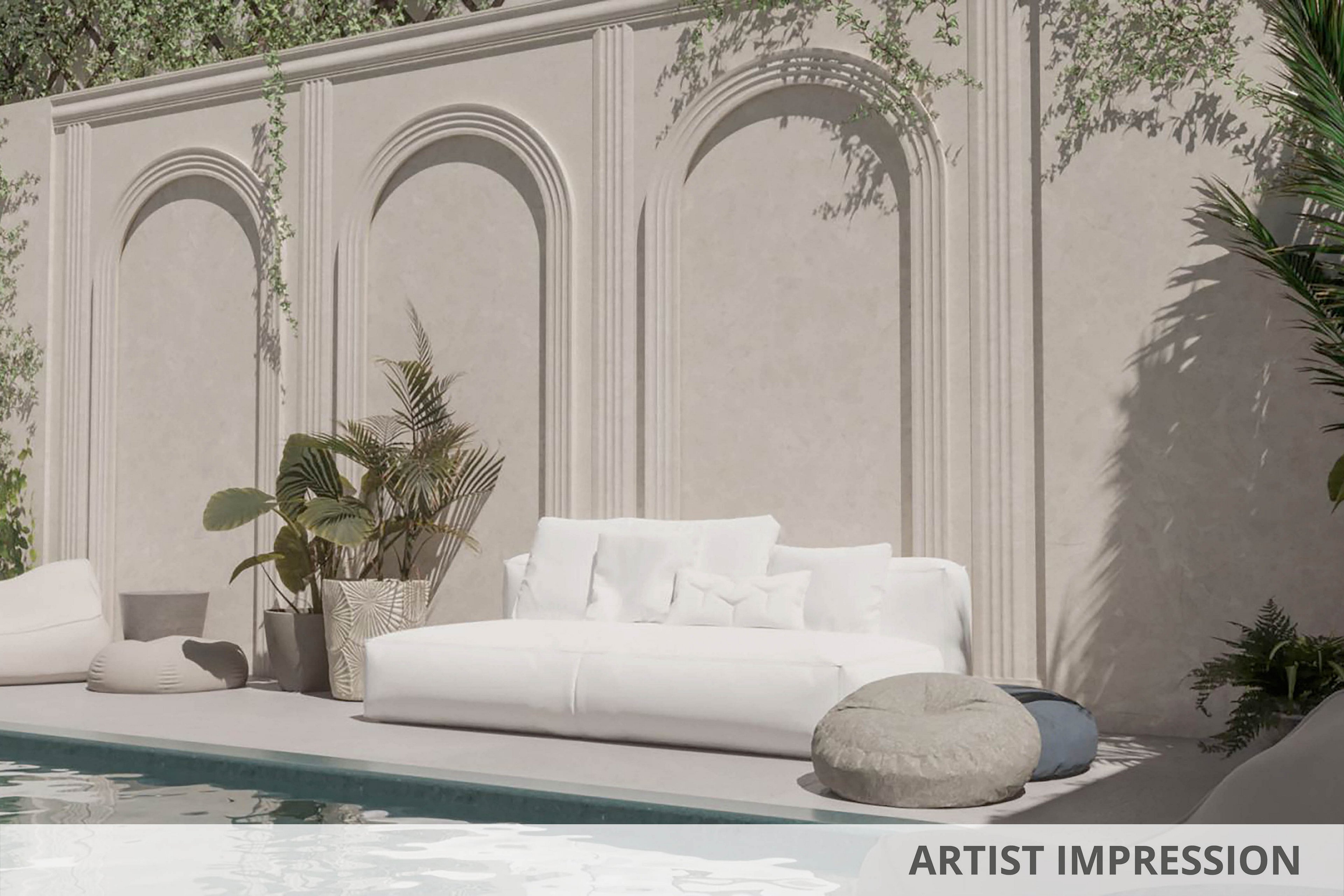 A contemporary outdoor seating area features a plush white sofa and cushioned poufs beside a serene swimming pool. The setting is framed by a decorative wall with arched niches and climbing greenery, creating a tranquil and stylish atmosphere. Neutral tones and soft textures dominate the scene, evoking a relaxed, minimalist vibe. Potted plants add a touch of nature to the elegant, sunlit space.
