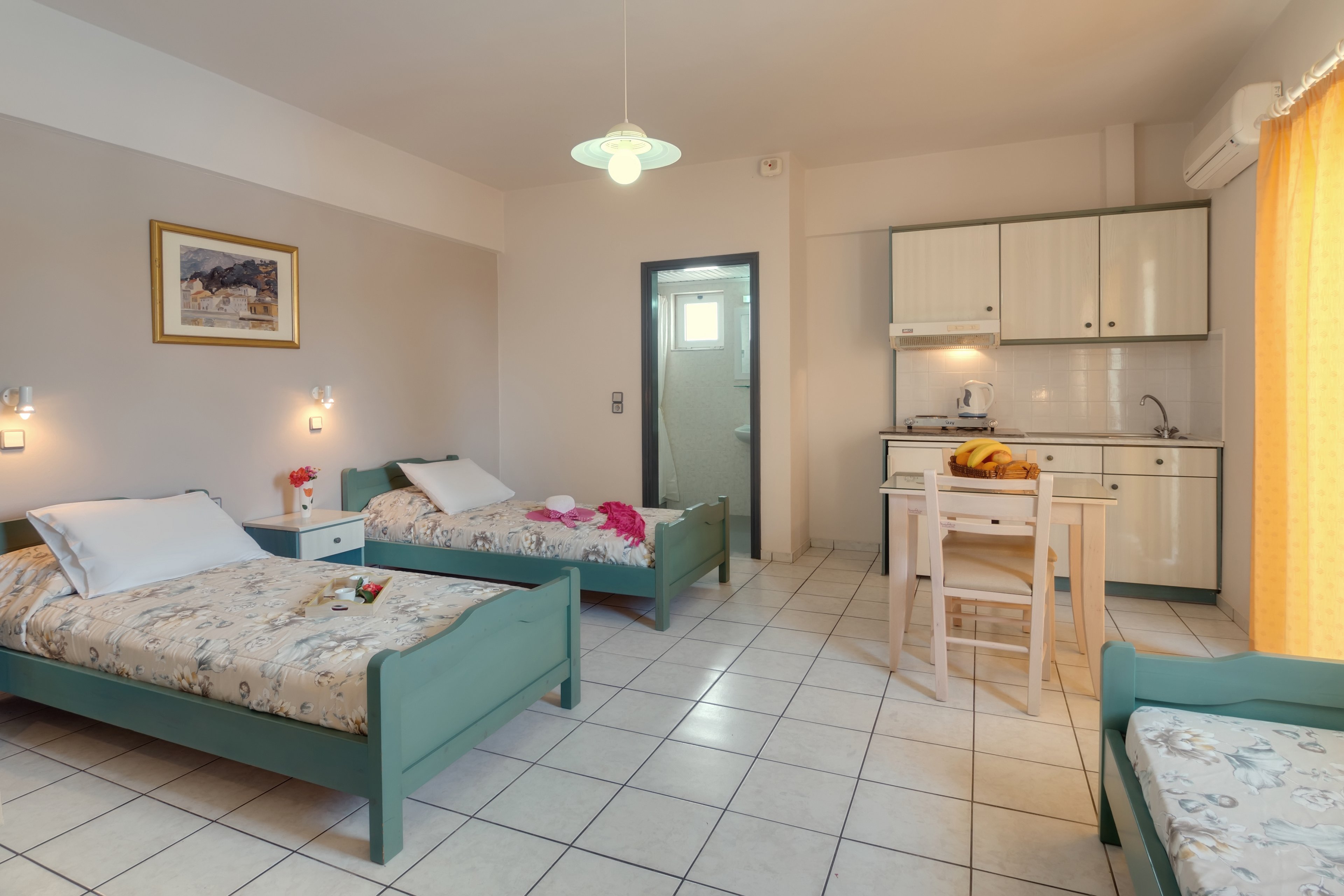 A tidy studio apartment features two single beds with floral bedding, a small dining table, and a compact kitchenette. The space is well-lit with natural light from a large window with yellow curtains. The room has tiled flooring, minimal decor, and a framed painting on the wall. The overall palette is soft and neutral, creating a calm and inviting atmosphere.
