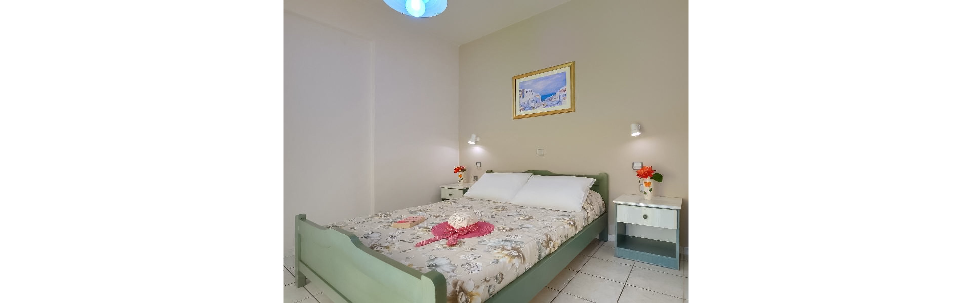 A neatly made double bed with a floral-patterned bedspread is the focal point of this simple hotel room. The setting features two bedside tables, each with a small vase of flowers, and a framed artwork above the bed. Soft lighting from wall-mounted lamps creates a welcoming atmosphere, while a straw hat and pink scarf rest on the bed. The room has a clean, minimalist style with light-colored walls and tiled flooring.