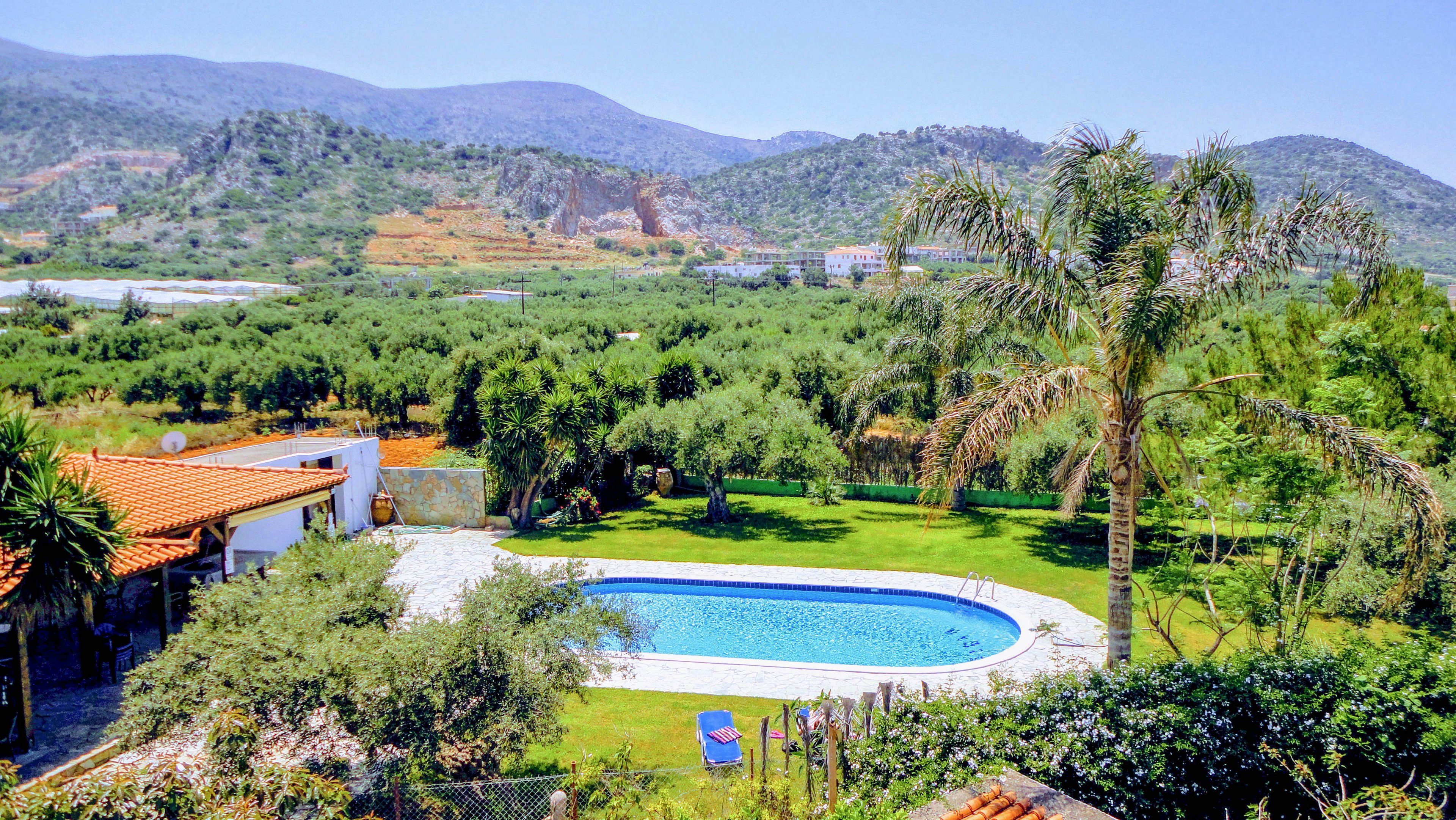 A private villa features a clear blue swimming pool surrounded by lush green grass and palm trees. The setting is a tranquil countryside with rolling hills and distant mountains under a bright, sunny sky. The scene evokes a relaxed, vacation mood with Mediterranean-style architecture and vibrant natural colors. No people or visible text are present in the image.