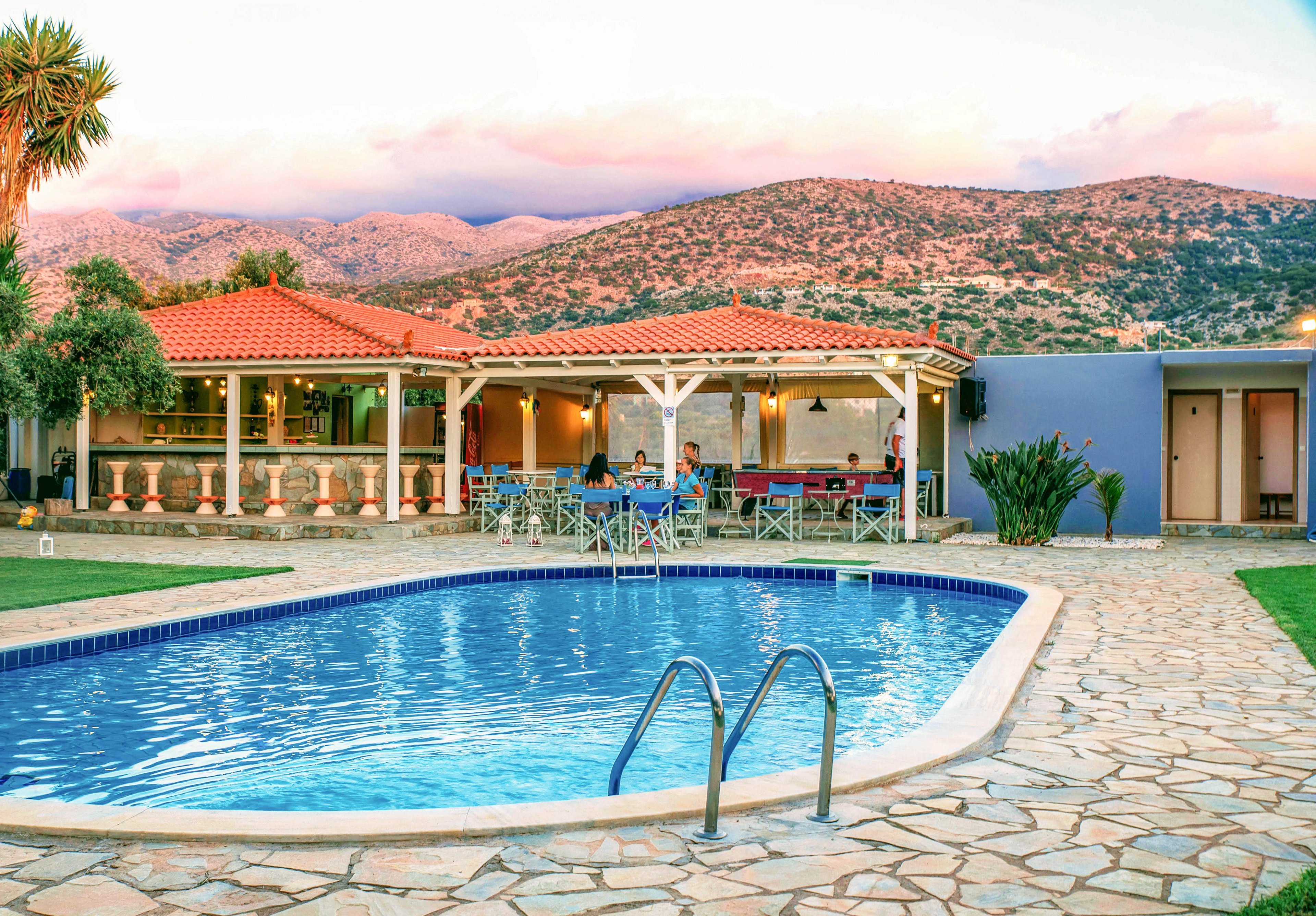 A serene outdoor pool is the focal point, surrounded by a stone patio and a covered dining area where several people are seated. The setting is a resort or villa with mountains in the background and a warm, inviting atmosphere. The sky is tinged with sunset colors, and the pool water reflects the tranquil ambiance. No visible text or numbers are present in the image.