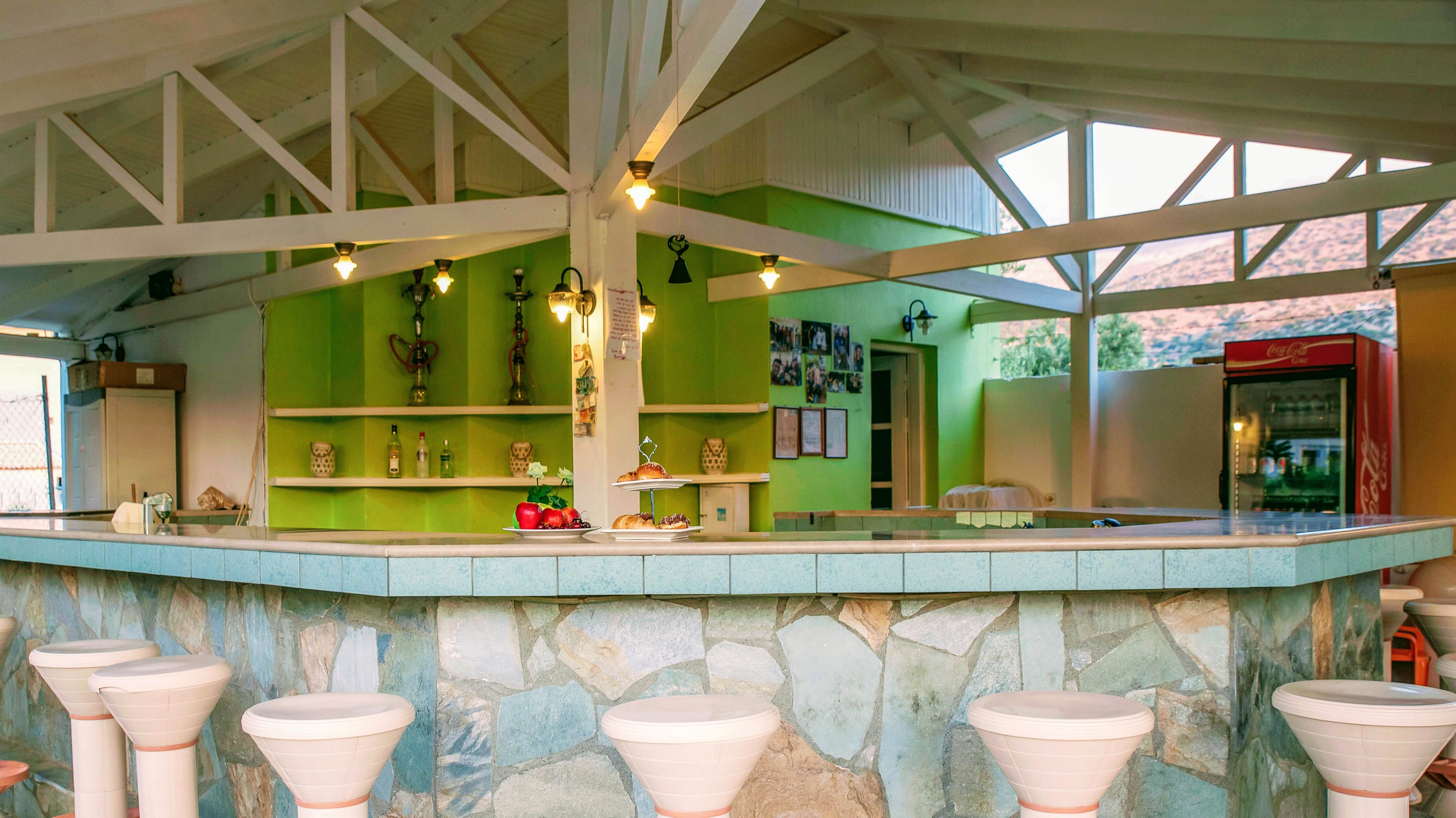 A spacious open-air bar features a stone-tiled counter and white bar stools arranged around it. The setting is bright and inviting, with green accent walls, shelves displaying bottles, and a refrigerator visible in the background. Overhead beams and soft lighting create a relaxed, tropical atmosphere. No people are present, and the area appears clean and ready for guests.