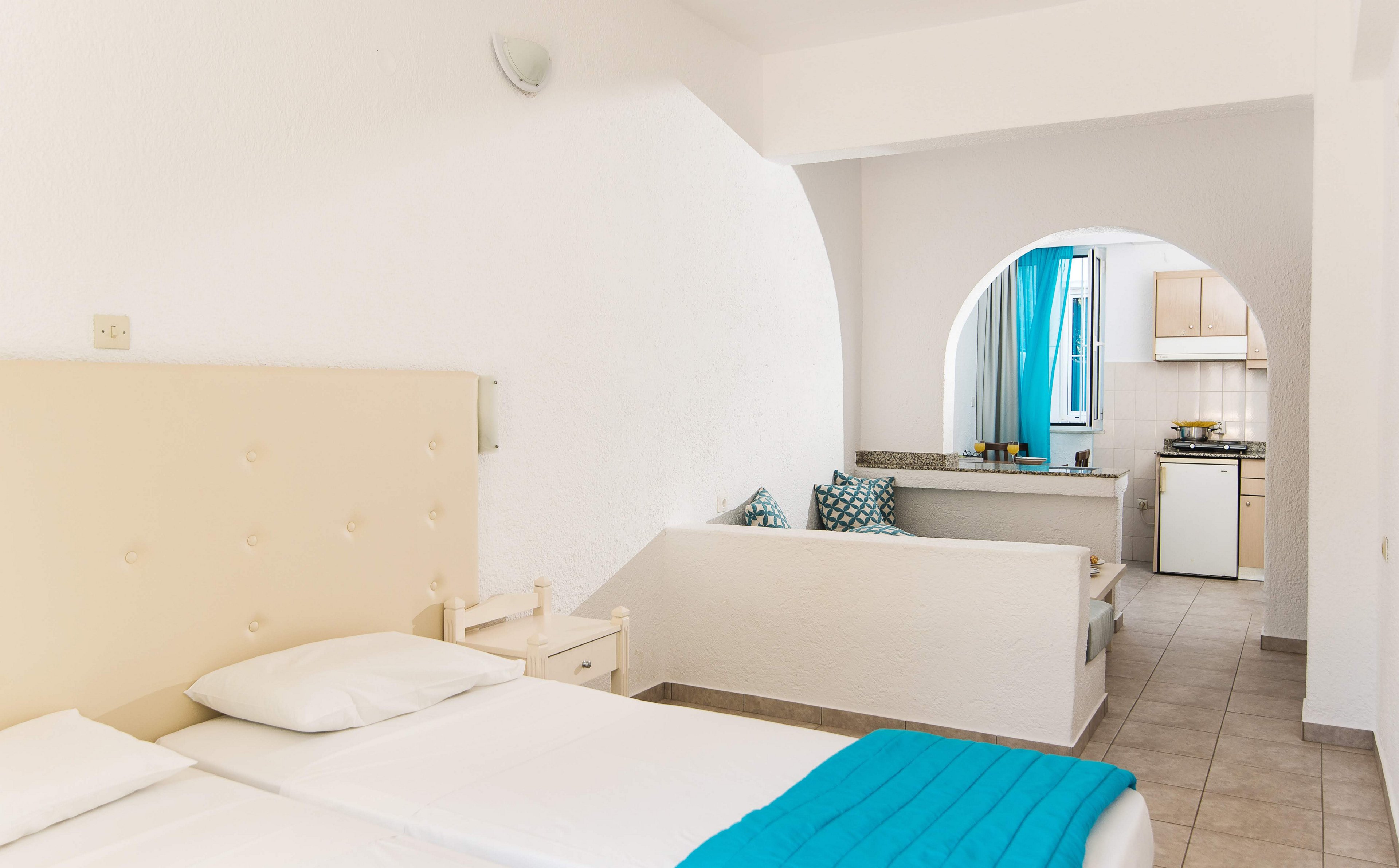 A spacious hotel room features two single beds with white linens and a turquoise throw. The room includes a small kitchenette in the background, separated by an archway, and a cozy seating area with patterned pillows. The decor is minimalist with a predominantly white palette and pops of blue, creating a fresh and inviting atmosphere. No visible text or numbers are present in the image.
