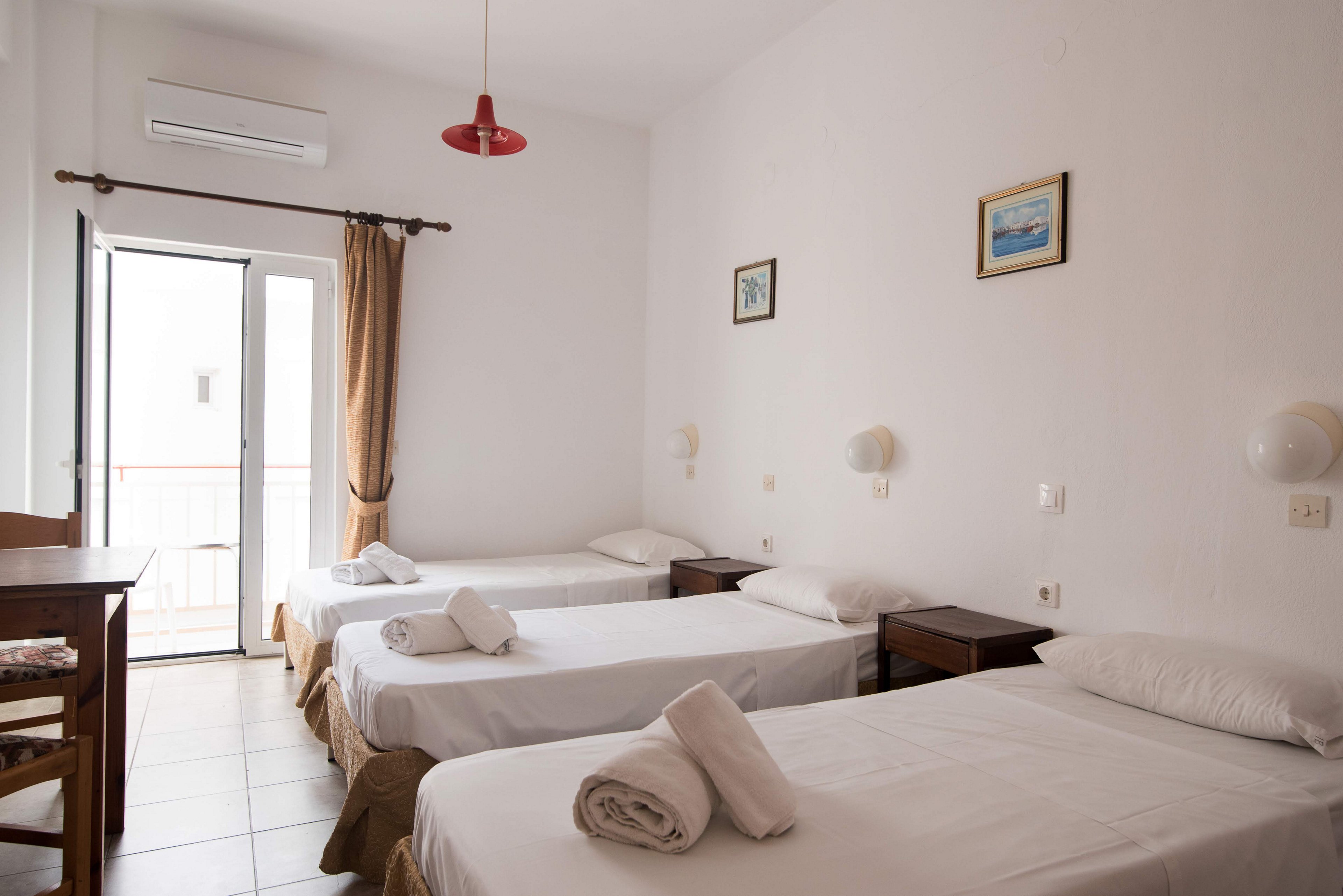 A clean, minimalist hotel room features three single beds neatly arranged with white linens and rolled towels. The space is well-lit by natural light from a balcony door, and simple decor includes framed art and a red ceiling lamp. Wooden furniture and tiled flooring add to the airy, uncluttered atmosphere, ideal for travelers or group stays.