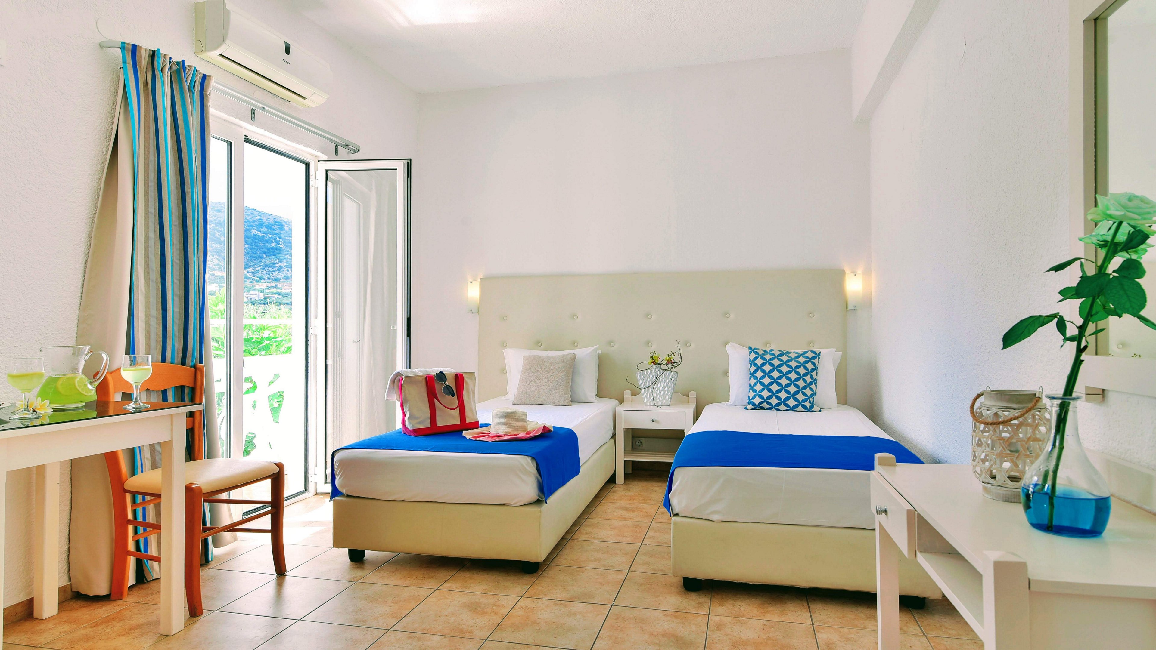 A modern hotel room features two single beds with blue accents and white linens, set against a clean, minimalist backdrop. The space is well-lit with natural light streaming in from a glass door leading to a balcony, offering a scenic outdoor view. A small table with drinks and a chair sits near the entrance, while a decorative vase with a flower adds a touch of freshness. The room's palette is dominated by white and blue, creating a calm and inviting atmosphere.