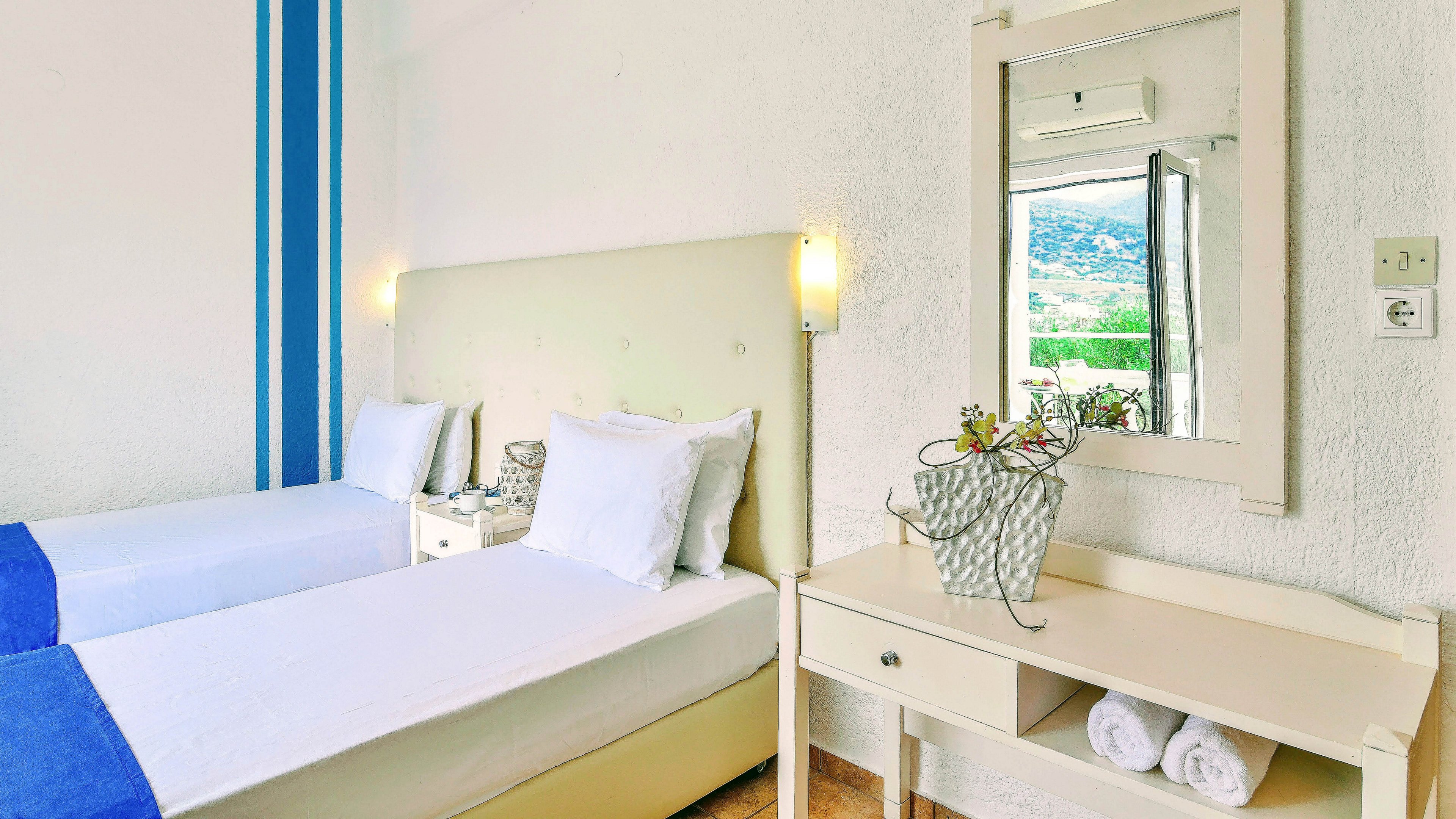 A clean, modern hotel room features two single beds with crisp white linens and blue throws. The room includes a light-colored headboard, a small desk with a mirror, and rolled towels neatly placed on a shelf. A vase with flowers adds a decorative touch, and sunlight streams in from a window with a scenic outdoor view. The overall palette is bright and airy, emphasizing relaxation and comfort.