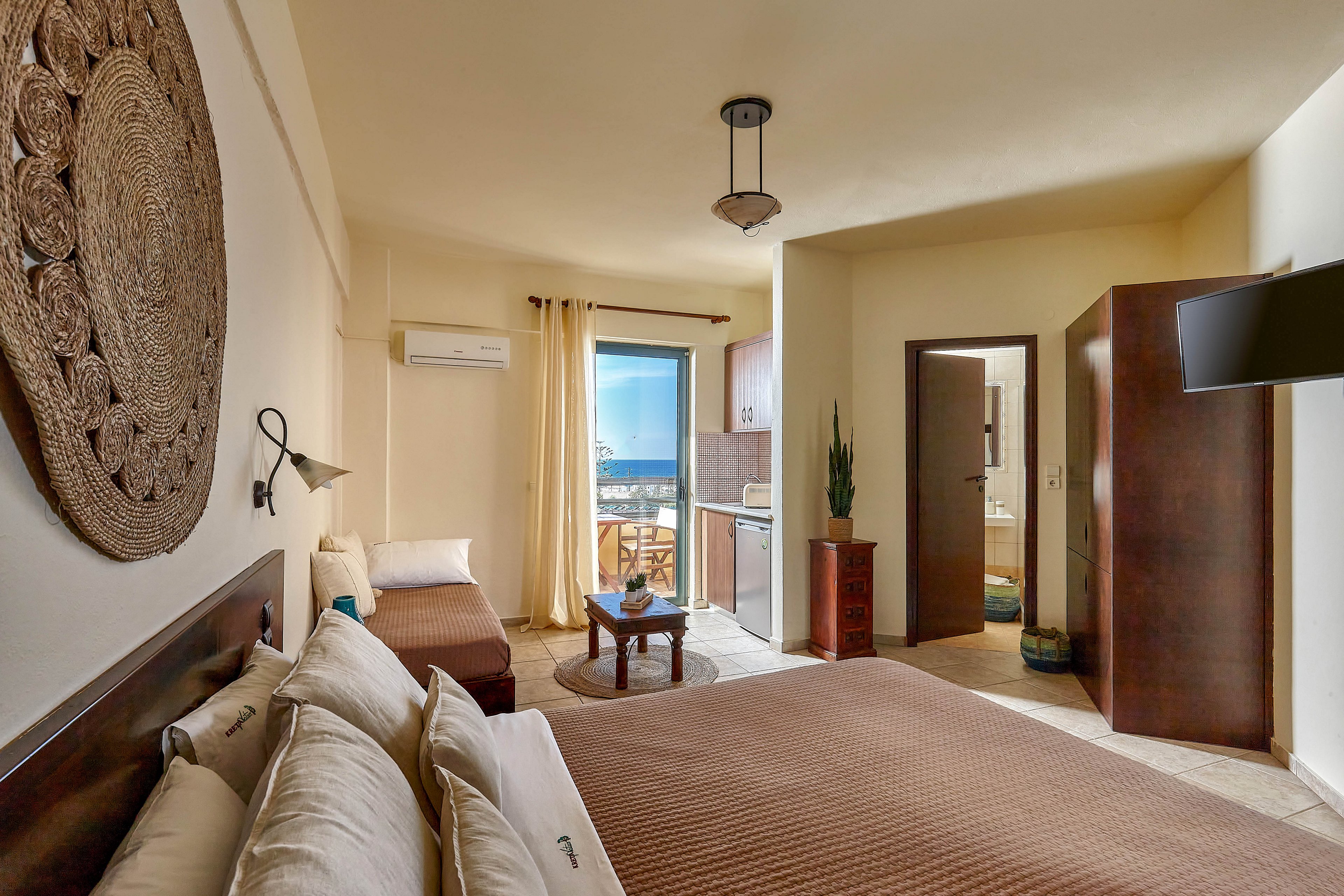 A spacious hotel room features a large bed, a single daybed, and a small table with chairs. The room opens to a balcony with a scenic sea view, letting in natural light through sheer curtains. Decor includes neutral tones, woven wall art, and minimalistic furnishings, creating a calm and inviting atmosphere.