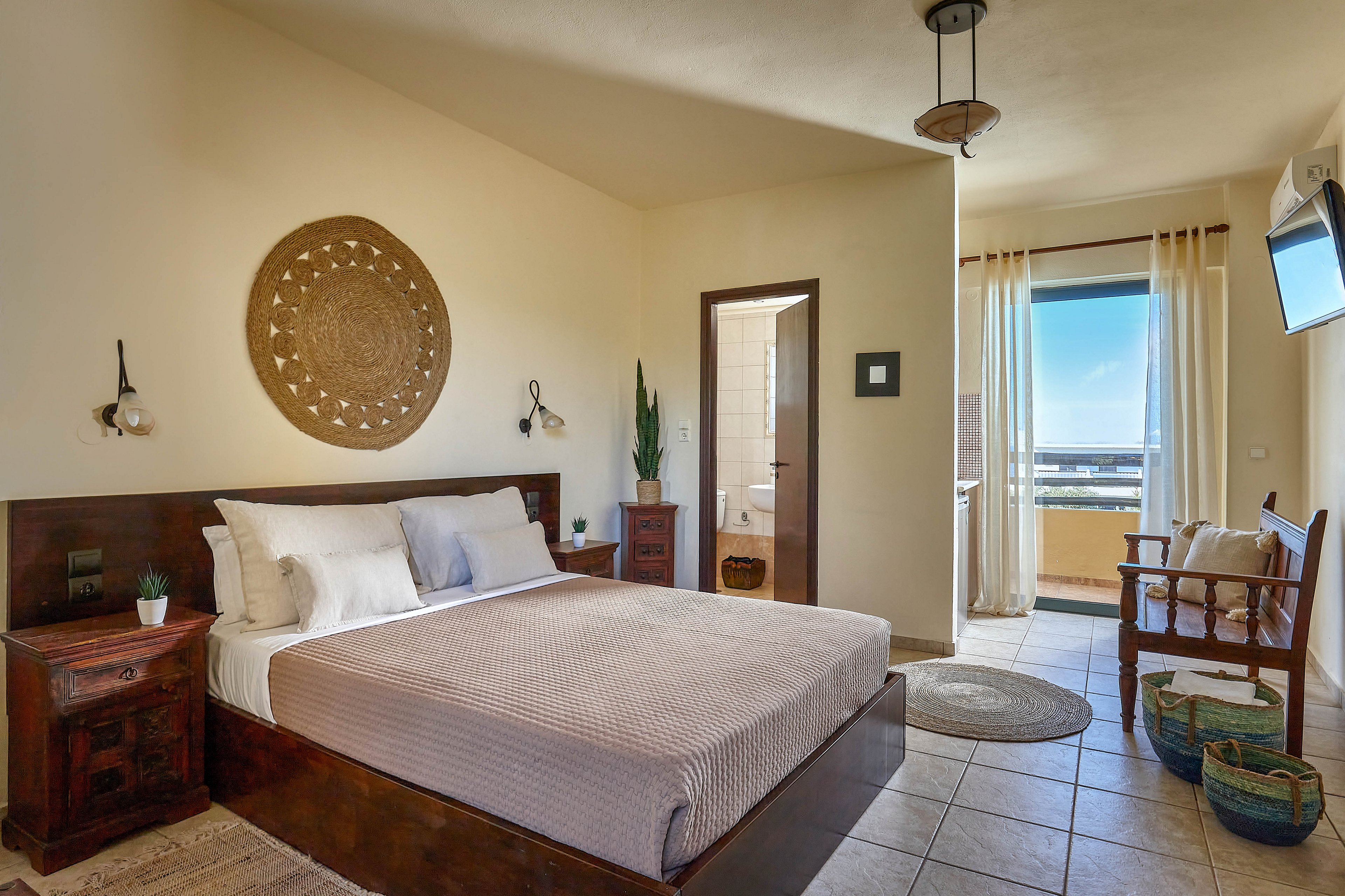 A spacious bedroom features a large bed with neutral bedding, wooden furniture, and a decorative woven wall piece above the headboard. The room is filled with natural light from a glass door leading to a balcony with a scenic view. Key visuals include tiled flooring, indoor plants, and woven baskets, creating a calm and inviting atmosphere.