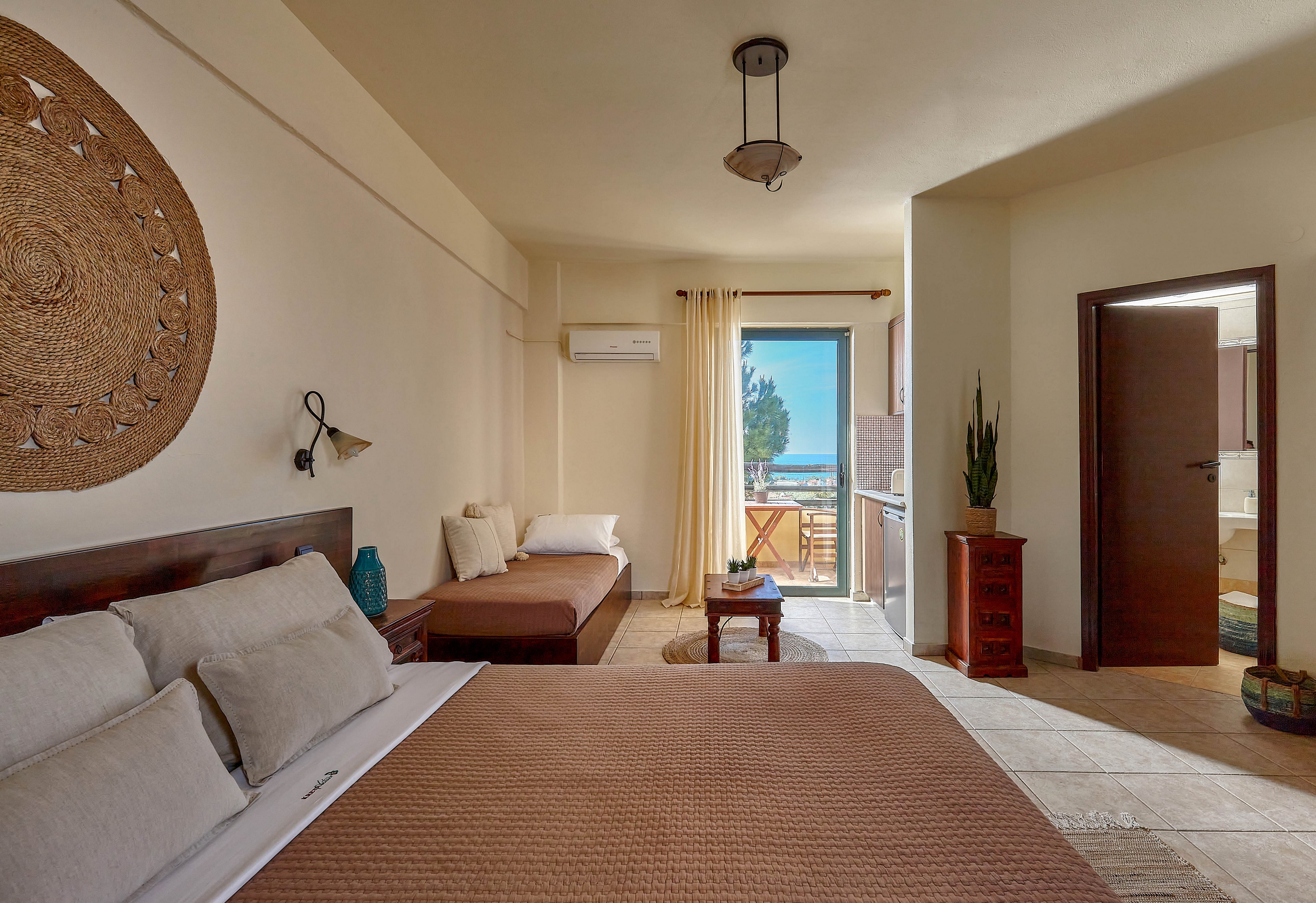 A neatly arranged hotel room features a large bed with brown bedding, a daybed, and a small table. The space is decorated in neutral tones with woven wall art and a potted plant, creating a calm and inviting atmosphere. Sunlight streams in through glass doors leading to a balcony with a view of the sea and trees. An air conditioning unit is mounted above the daybed, and the room is well-lit with natural light.