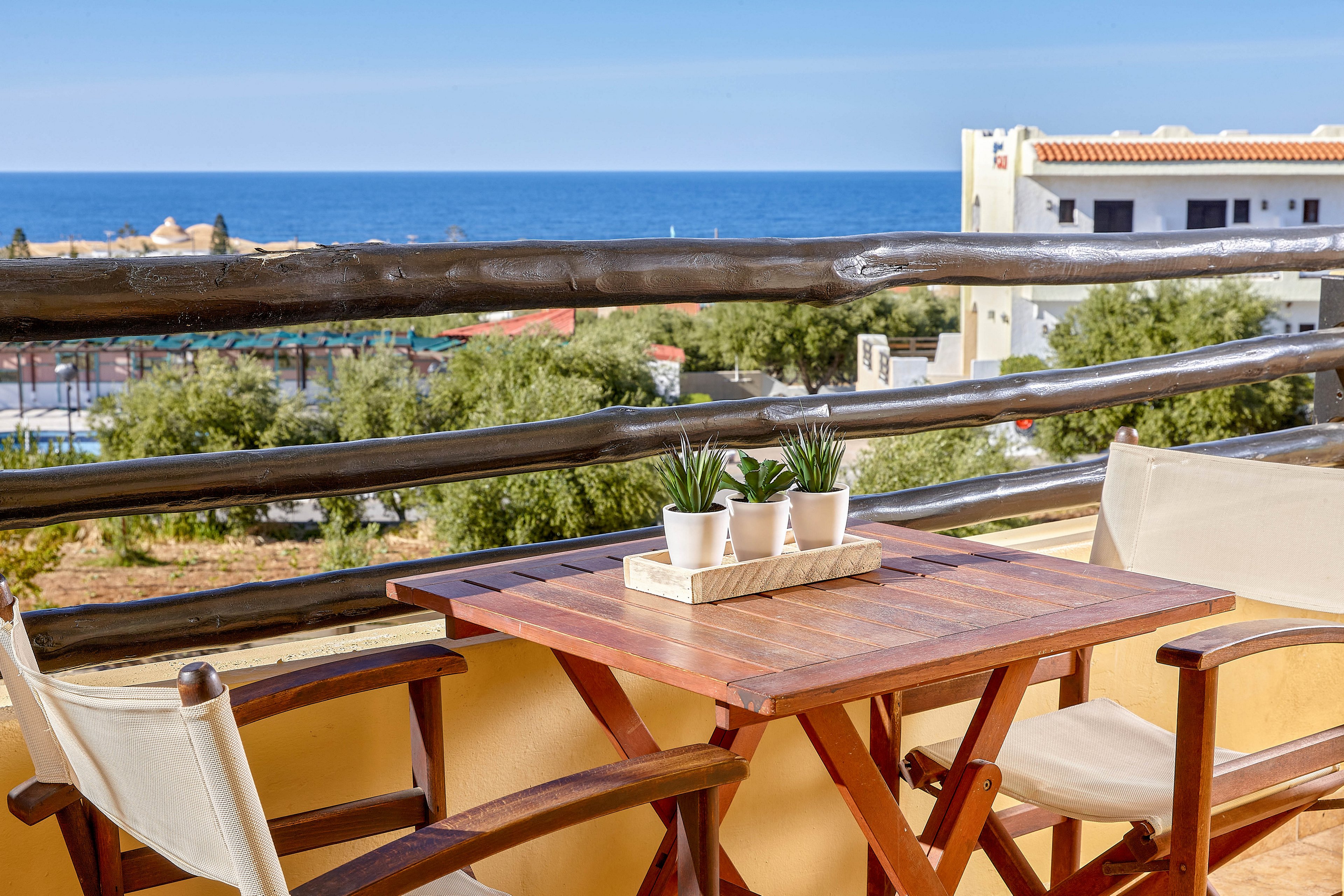 A wooden table with two chairs is set on a balcony overlooking the ocean. The table features a tray with four small potted plants, adding a touch of greenery to the scene. The background includes a clear blue sky, distant buildings, and lush trees, creating a relaxing and inviting atmosphere. The overall mood is bright and tranquil, ideal for leisure or vacation settings.