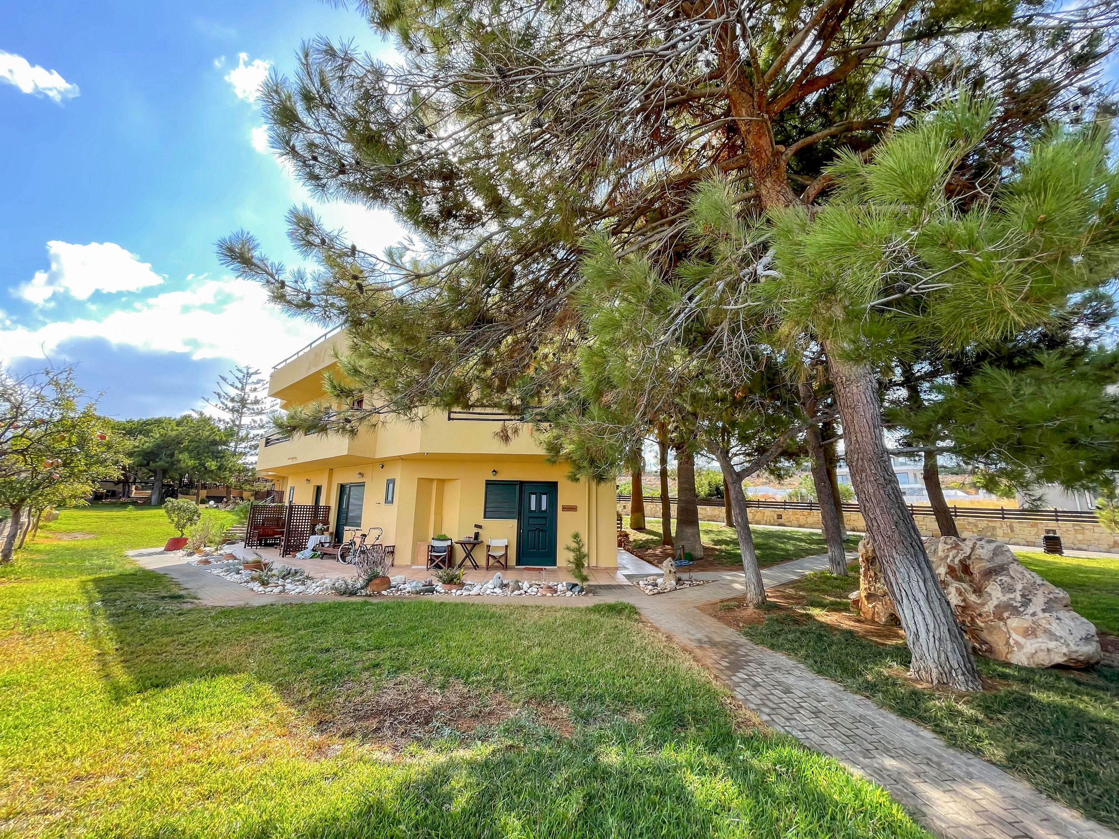 A two-story yellow house stands in a spacious, grassy yard with a paved walkway leading to the entrance. The patio area features outdoor seating and is shaded by tall pine trees, creating a relaxed and inviting atmosphere. The scene is brightly lit with clear blue skies and scattered clouds, emphasizing a peaceful suburban or rural setting.