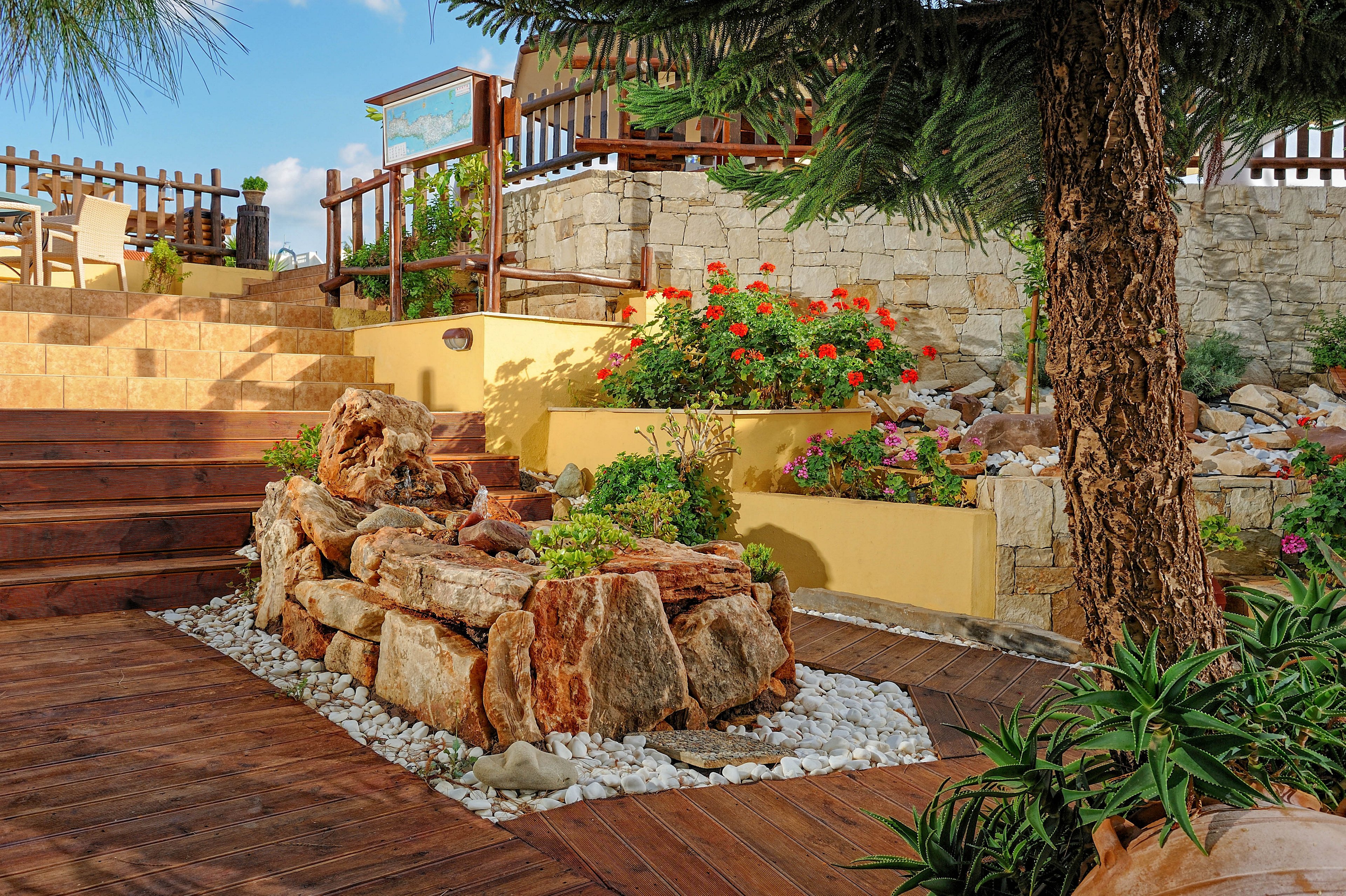 A vibrant outdoor terrace garden features a central stone planter filled with greenery and surrounded by white decorative rocks. The setting includes wooden decking, colorful flowers, and a rustic stone wall in the background. Bright sunlight enhances the cheerful mood, with seating visible on the upper level. The scene is lush, inviting, and well-maintained, ideal for relaxation or outdoor gatherings.