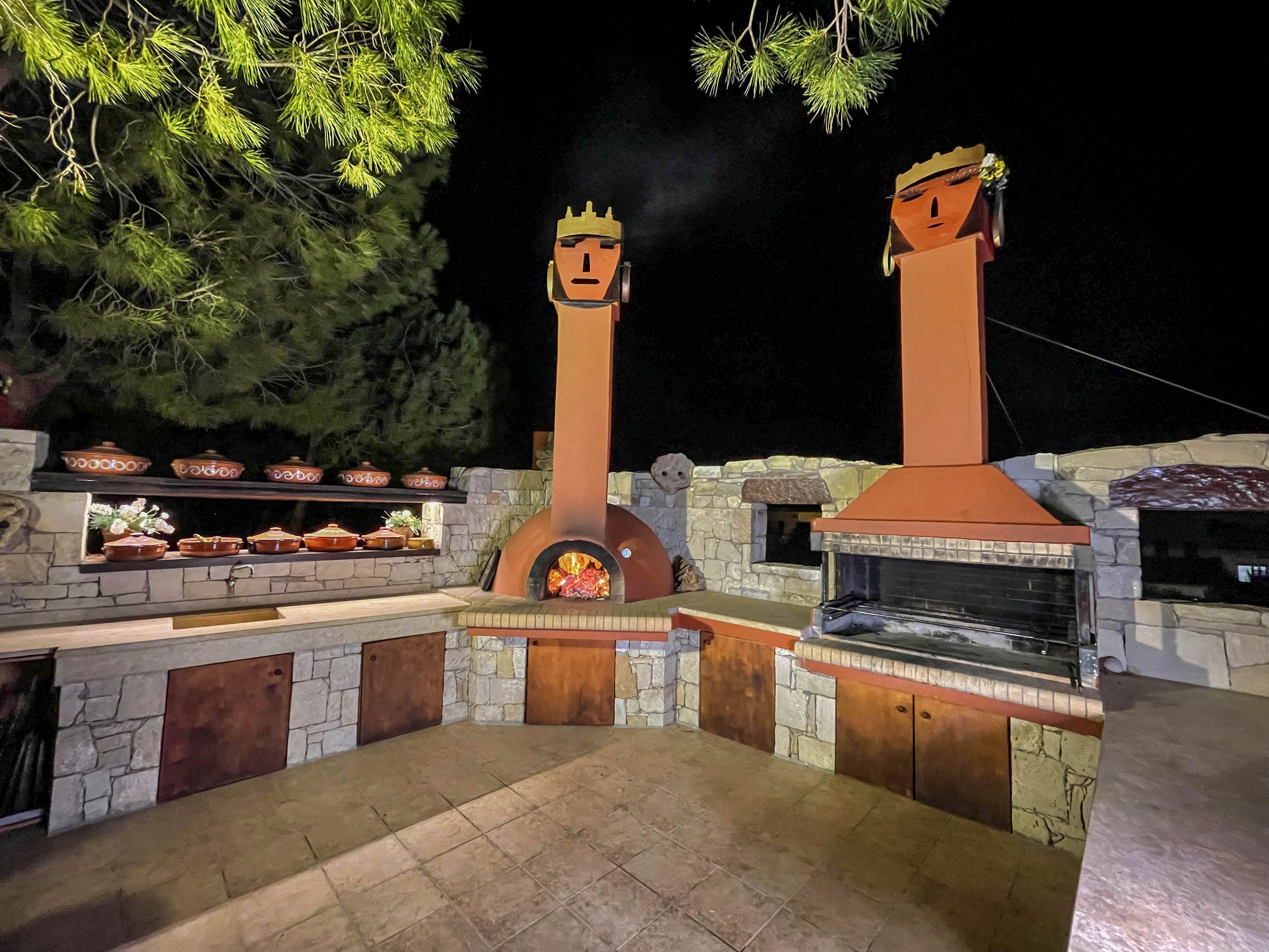 A spacious outdoor kitchen features stone countertops and cabinetry, highlighted by two tall chimneys with stylized face designs. The setting is illuminated at night, with a wood-fired oven glowing and various clay pots arranged on the counter. Pine tree branches frame the scene, adding a natural touch to the rustic ambiance.