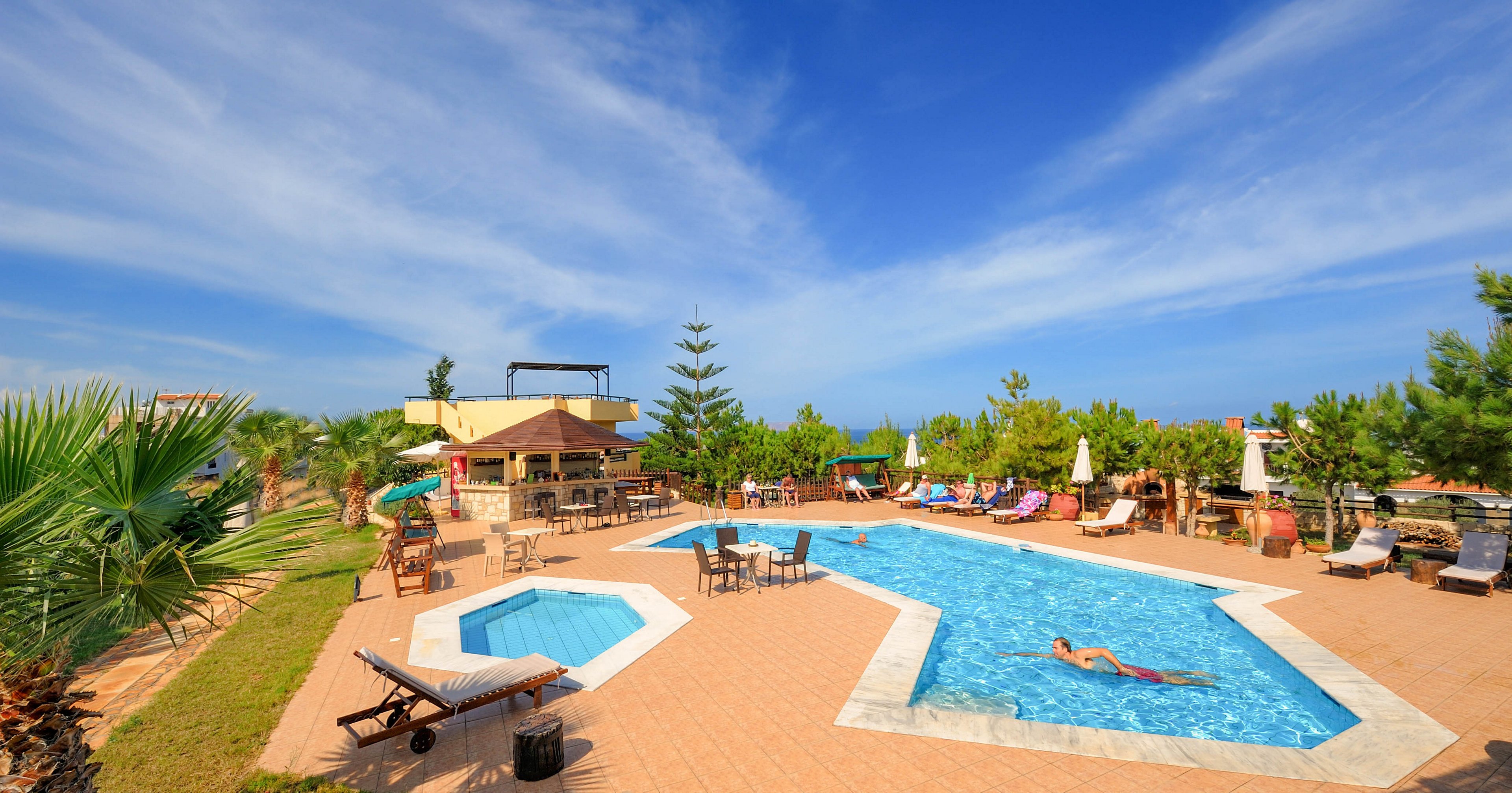 A spacious outdoor swimming pool is the main focus, surrounded by lounge chairs and tables under a bright blue sky. A single adult is relaxing in the pool, while the area is bordered by lush greenery and palm trees. The setting suggests a tranquil, vacation-like atmosphere with clear weather and vibrant colors. No visible text or numbers are present in the image.