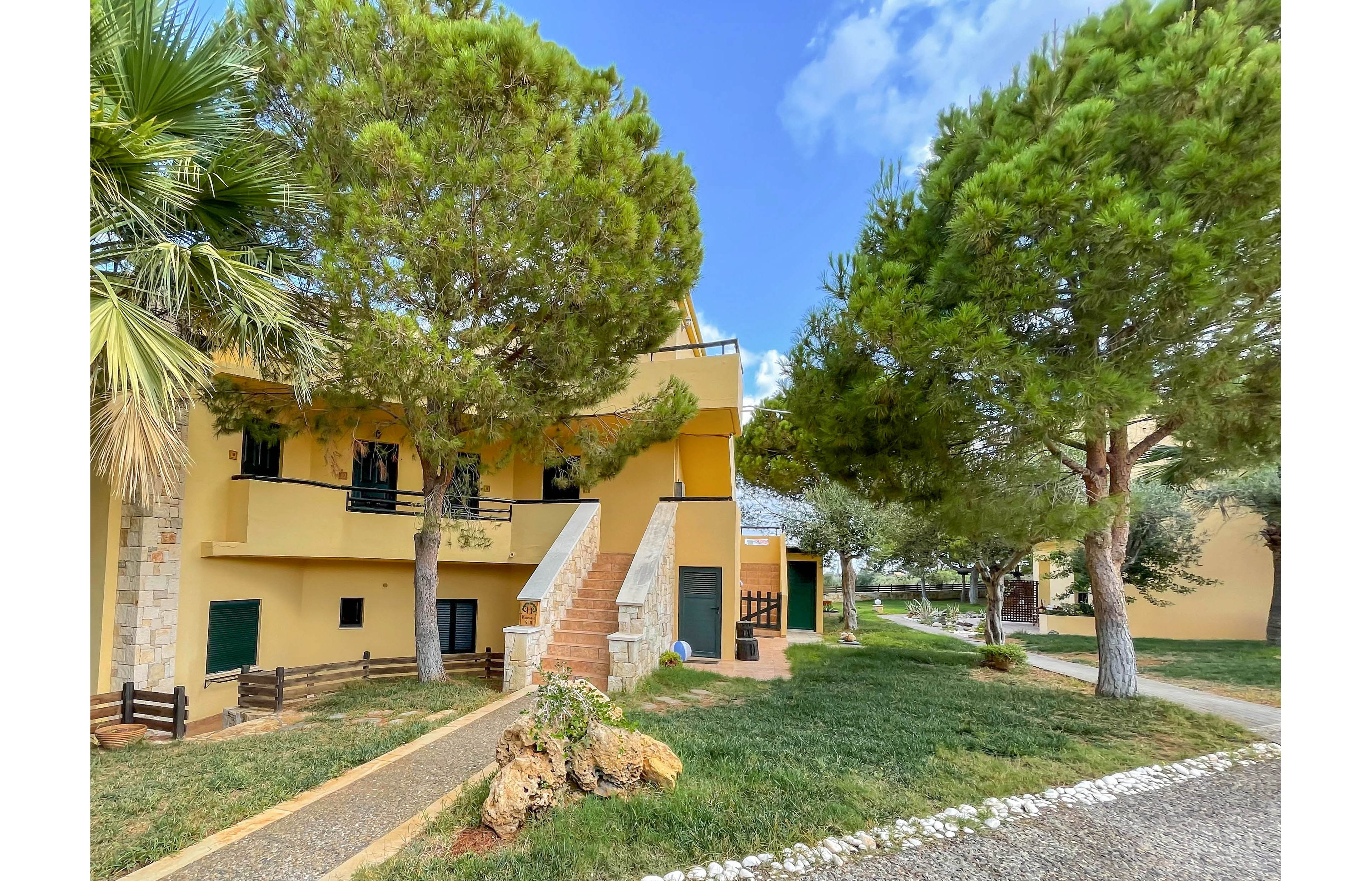 A two-story yellow villa is surrounded by tall, leafy green trees and a well-kept grassy yard. The building features stone accents, exterior staircases, and multiple balconies with black railings. The setting is peaceful and sunlit, with a clear blue sky and a stone pathway leading to the entrance. No visible people, text, or numbers are present in the image.