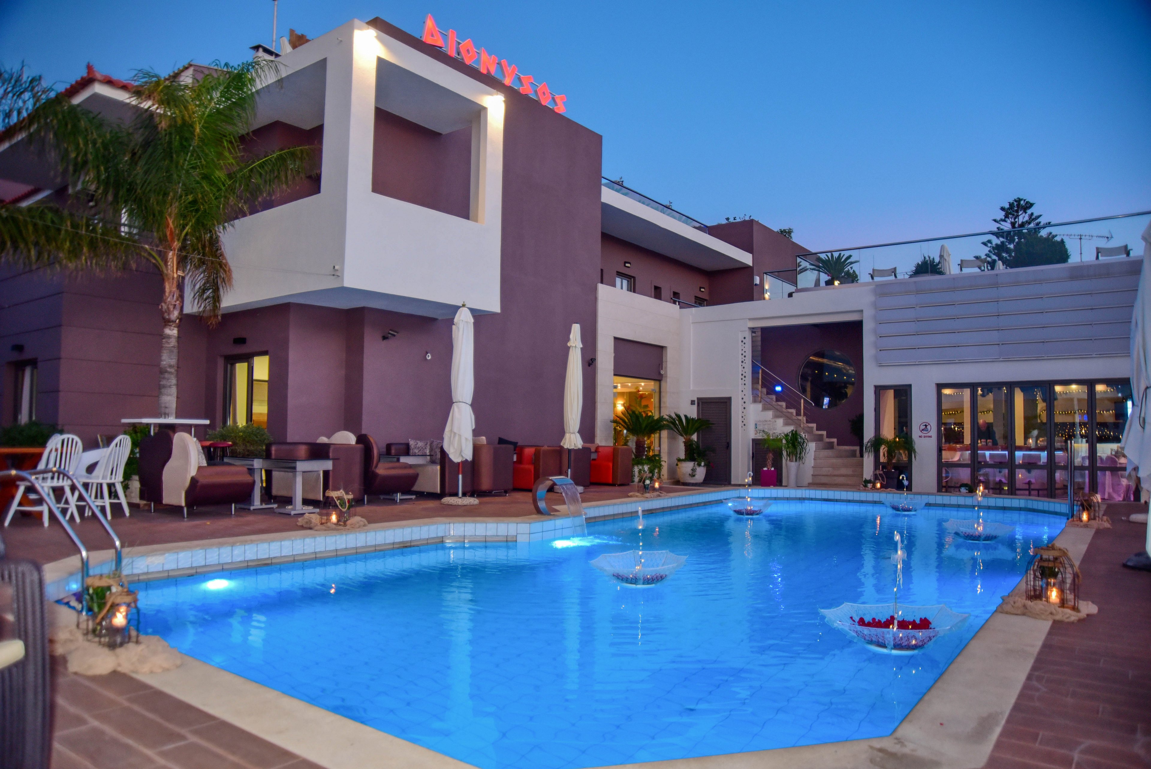 A contemporary hotel building features a large, illuminated outdoor swimming pool in the foreground. The setting is at dusk, with clear blue skies and soft lighting creating a relaxing atmosphere. Poolside seating, umbrellas, and decorative floating trays are visible, enhancing the inviting ambiance. The hotel name is clearly displayed in red letters on the rooftop.