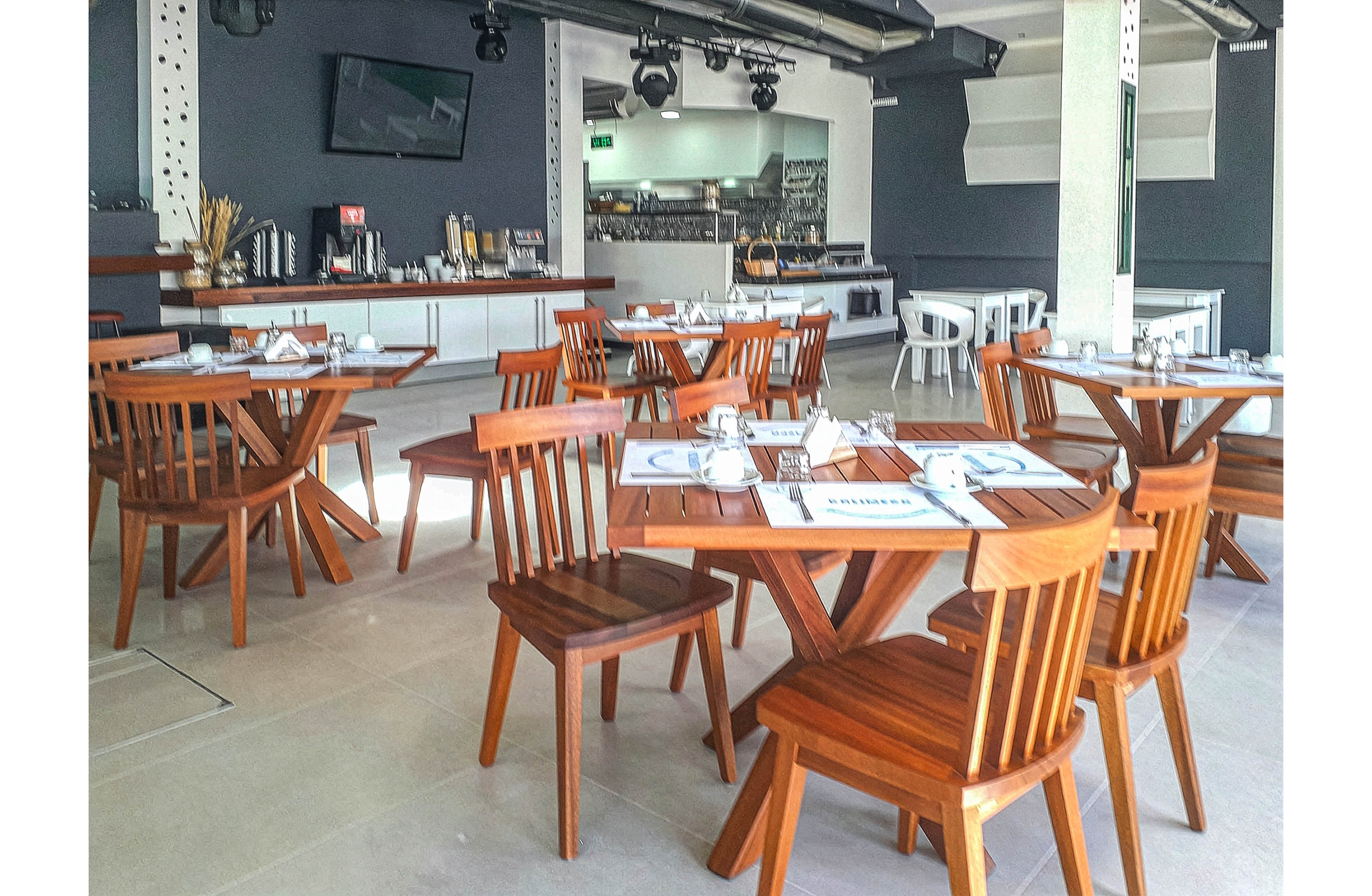 A contemporary restaurant dining area features several wooden tables and chairs arranged neatly. The setting is bright with natural light, and the decor is minimalist with clean lines and neutral tones. Place settings are visible on the tables, and a buffet or service counter is seen in the background. The overall atmosphere is calm and inviting, suitable for casual dining. No visible text or numeric values are present in the image.