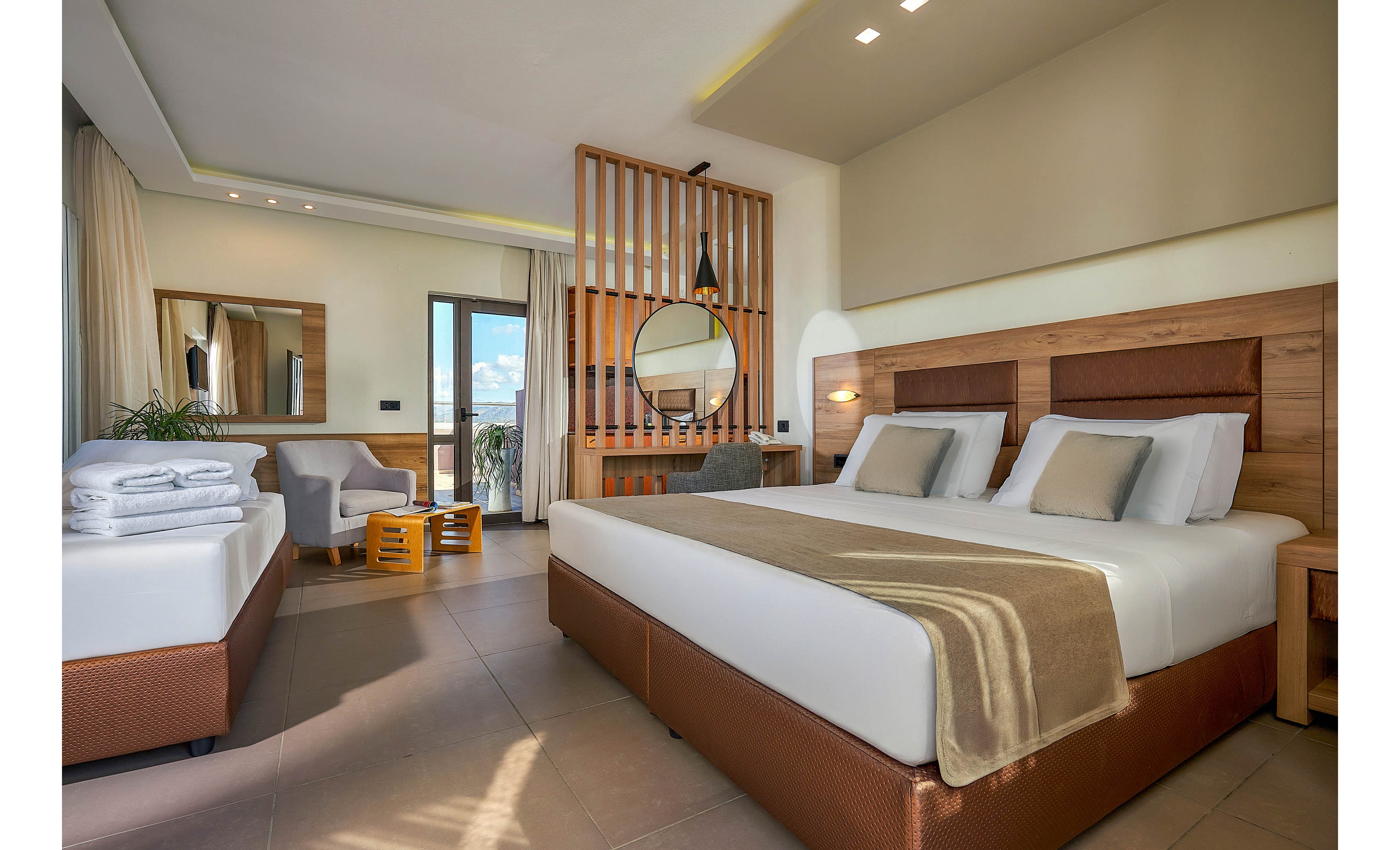 A spacious, contemporary hotel room features a large double bed with neutral bedding and a single bed neatly arranged with towels. The room includes a seating area, wooden accents, and a round mirror on a slatted divider. Natural light fills the space through a glass door leading to a balcony, creating a bright and inviting atmosphere.