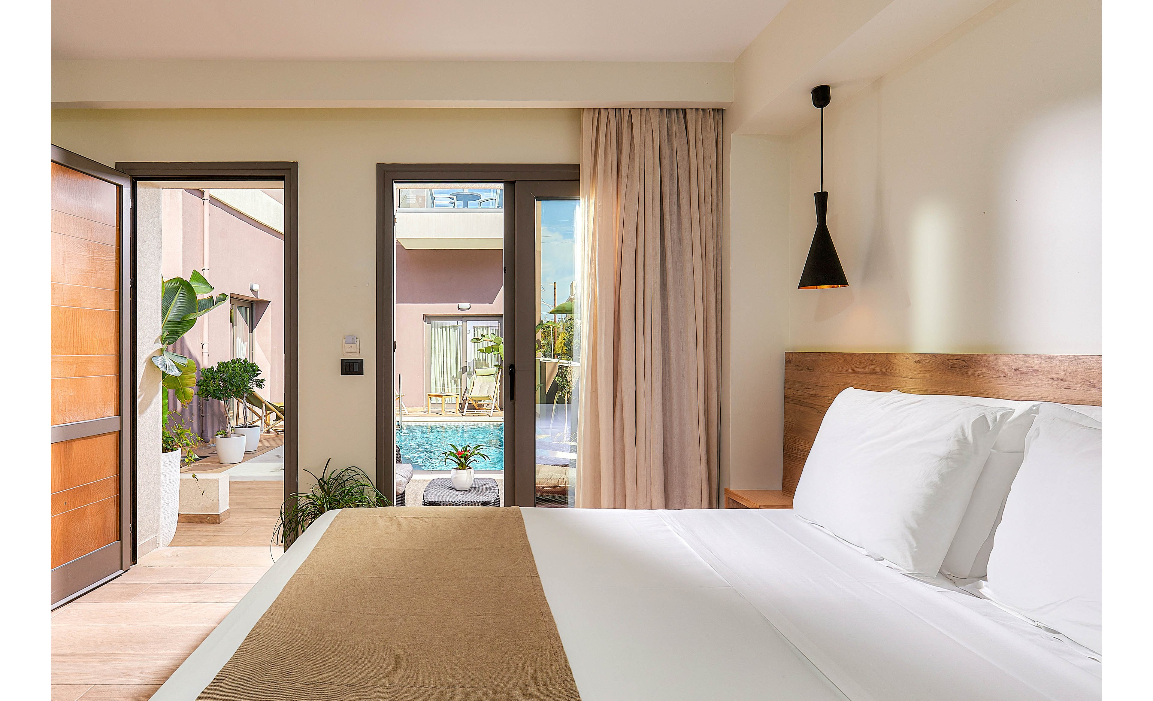 A bright, contemporary hotel room features a neatly made bed with white linens and a tan throw. Large glass doors open to a private patio with a small pool, potted plants, and outdoor seating. The room is decorated in neutral tones with minimalist lighting and natural sunlight streaming in, creating a relaxing atmosphere.