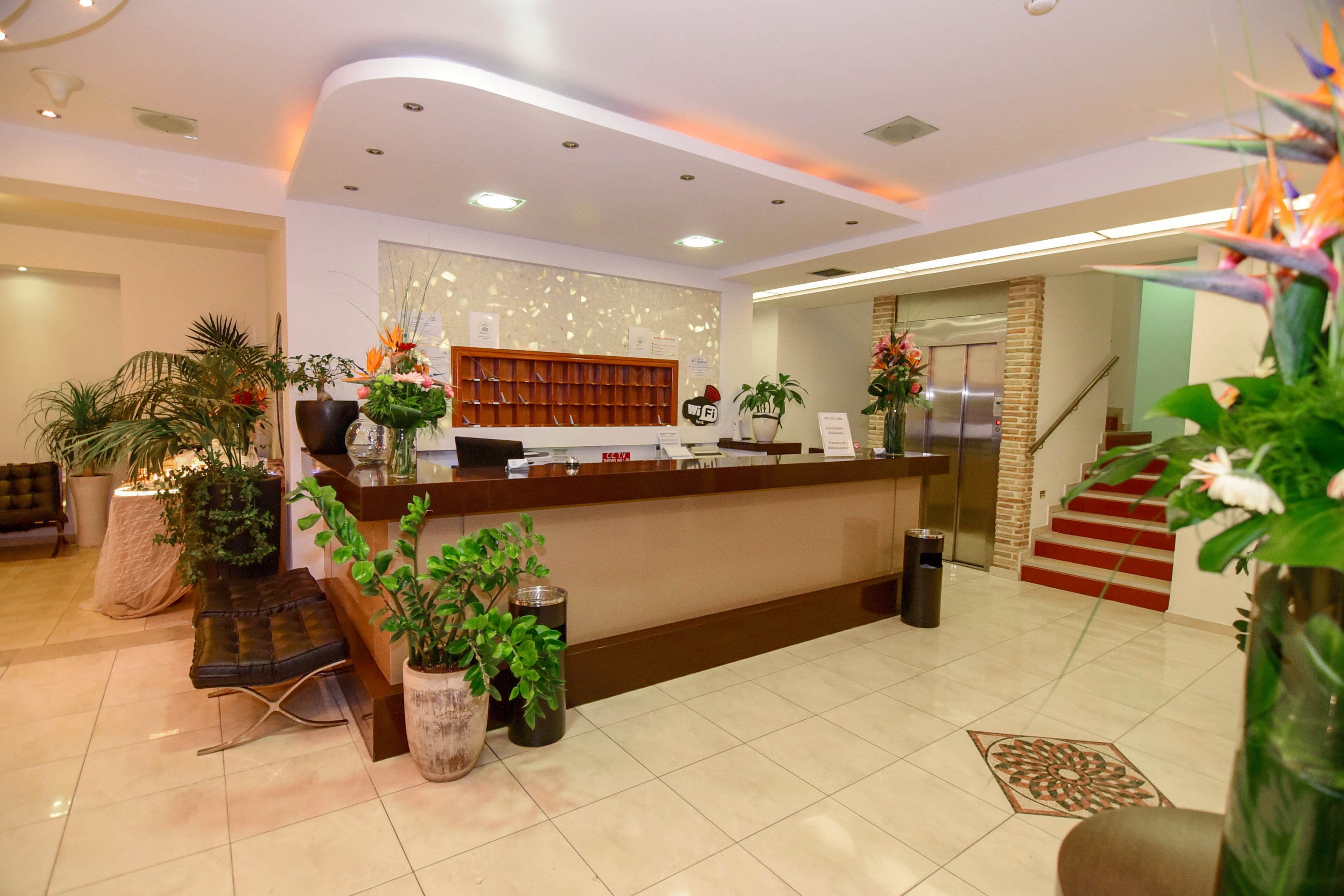 A contemporary hotel reception desk is the main focus, surrounded by lush indoor plants and elegant decor. The setting features a tiled floor, a staircase with red carpeting, and a well-lit ceiling with recessed lighting. Decorative vases and a patterned floor tile add visual interest, while the mail slots behind the desk suggest a hospitality environment. No visible text or numbers are present in the image.