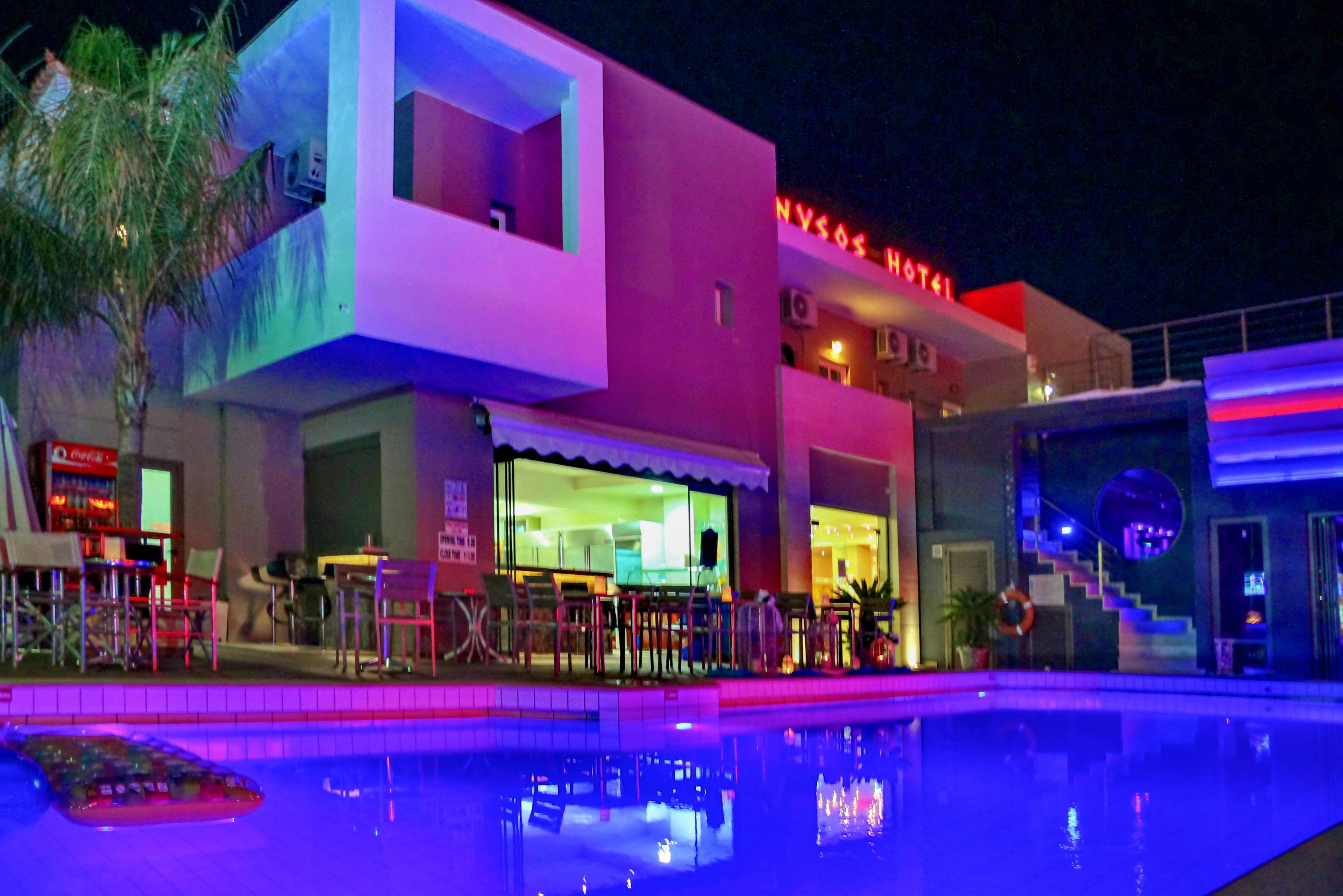 A contemporary hotel building is shown at night, featuring a brightly lit swimming pool in the foreground. The scene includes outdoor seating areas, neon lighting, and visible architectural details. The word 'NYSSOS HOTEL' is clearly illuminated on the building, adding to the vibrant nighttime atmosphere.