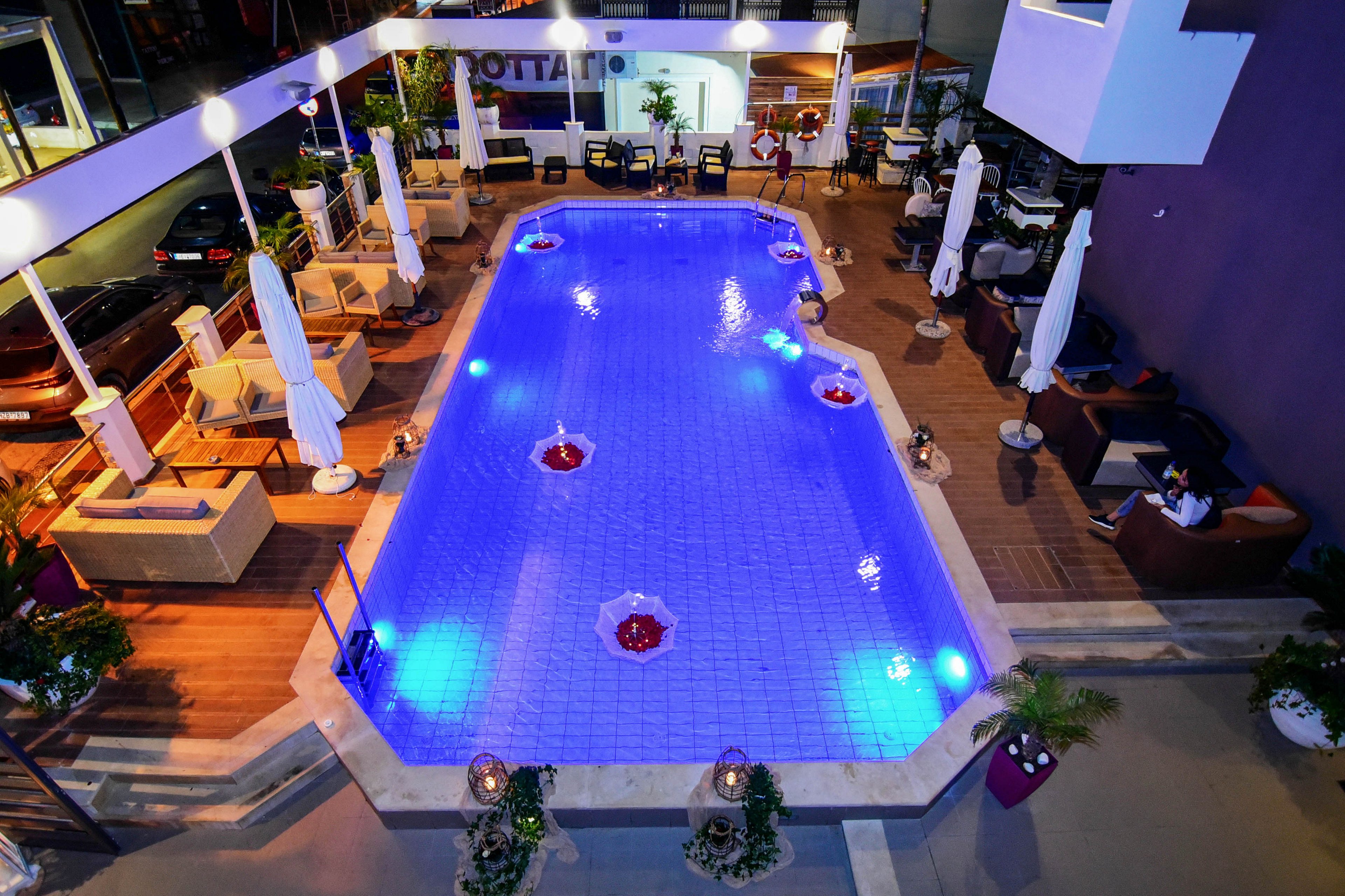 A rectangular swimming pool is illuminated with blue lights, creating a vibrant nighttime atmosphere. Surrounding the pool are lounge chairs, tables, and large white umbrellas, with potted plants adding greenery to the scene. The setting appears to be a hotel or upscale residential complex, with a relaxed and inviting mood. The pool deck is made of wood and tile, and the overall palette features cool blues and warm neutrals. No visible people or explicit numeric values are present in the image.