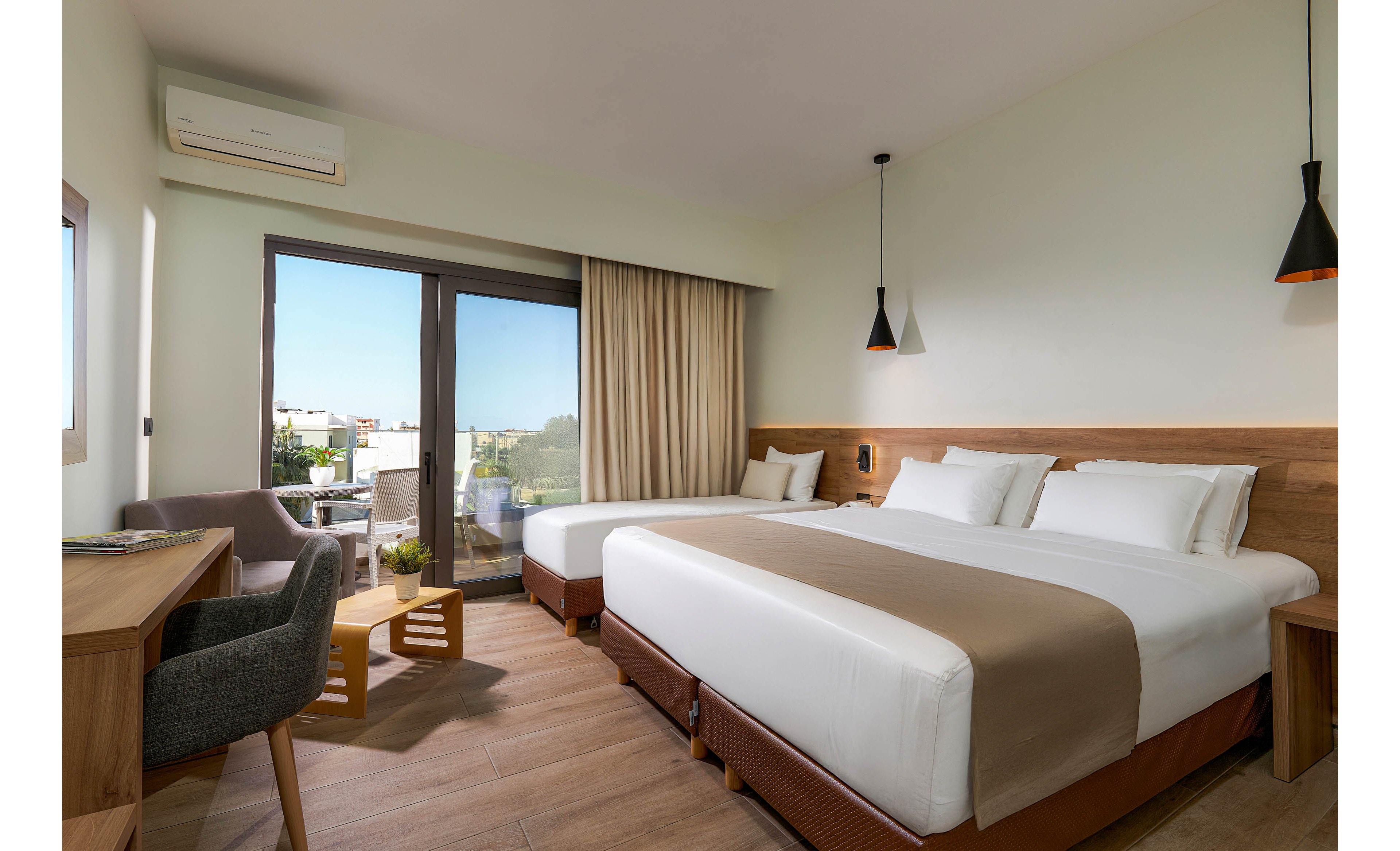 A contemporary hotel room features two large beds with white linens and beige runners. The space includes a wooden desk, a small sitting area with chairs and a table, and a sliding glass door leading to a balcony with an outdoor view. The room is decorated in neutral tones with minimalist lighting and natural light streaming in. An air conditioning unit is visible above the window.