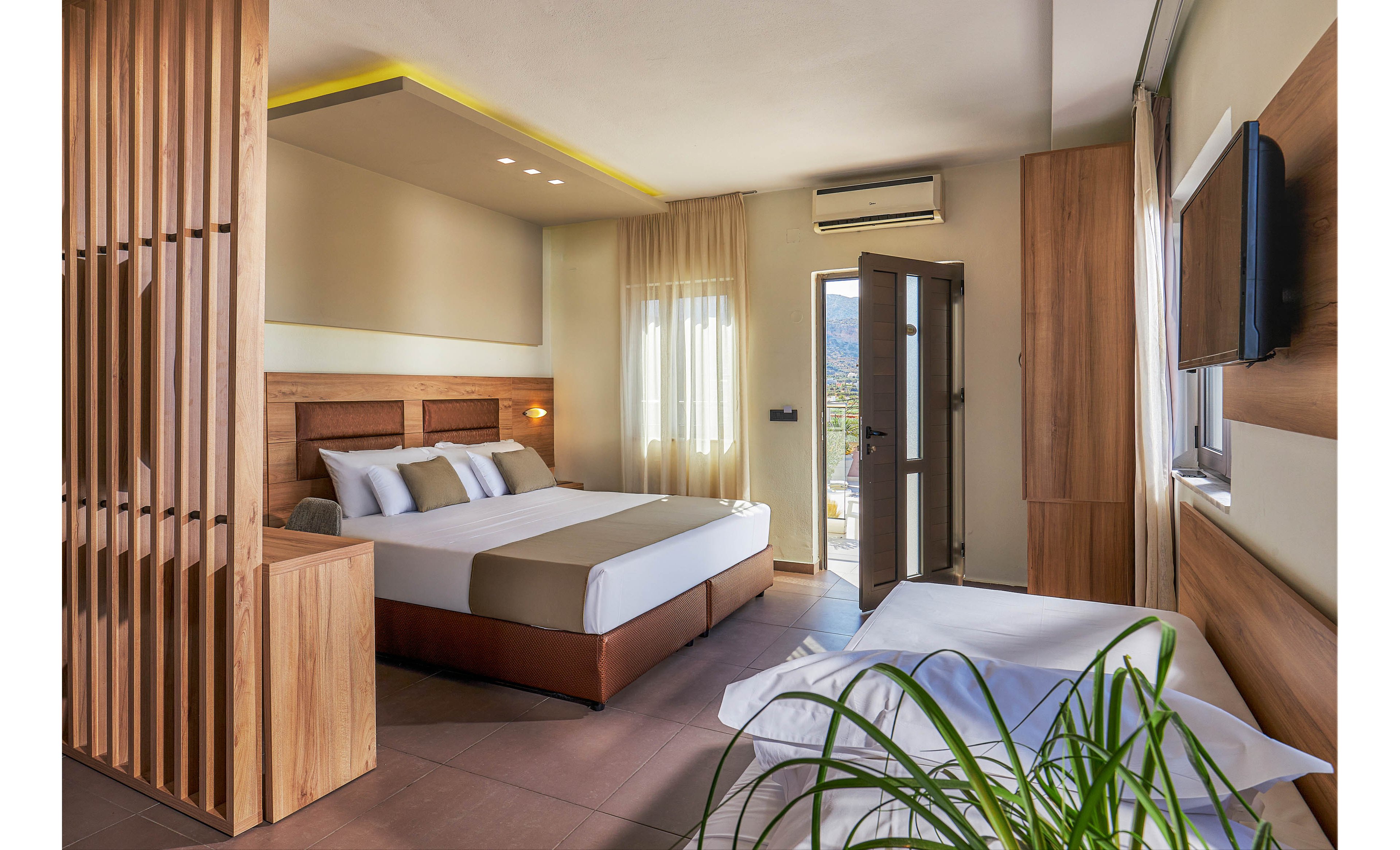 A contemporary hotel room features a large double bed with crisp white linens and accent pillows. The space is decorated in neutral tones with wooden accents, a wall-mounted television, and a visible air conditioning unit above the door. Natural light streams in through sheer curtains, and a small balcony is accessible through a glass door. A green plant adds a touch of freshness to the clean, minimalist design.