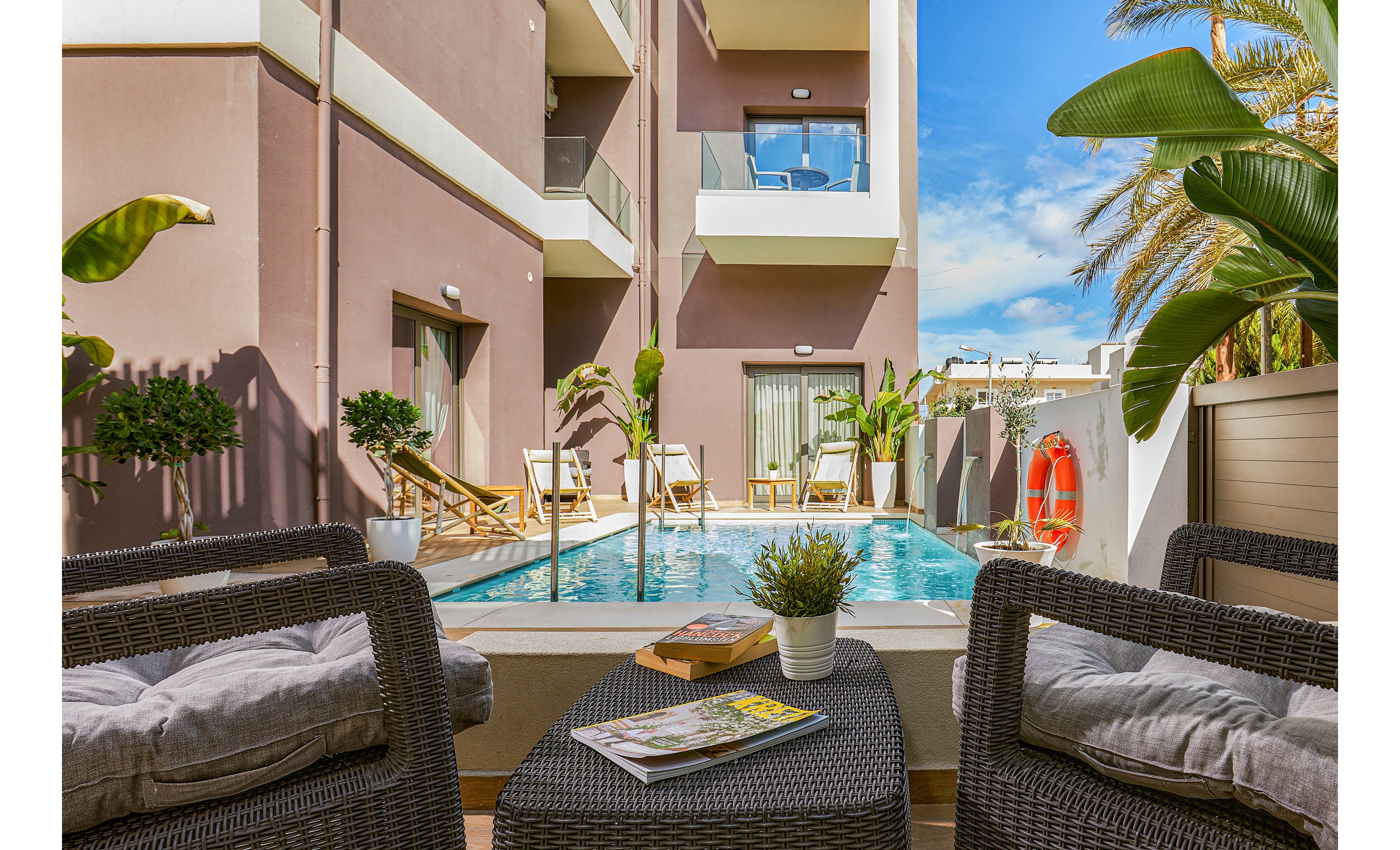 A contemporary hotel courtyard features a small swimming pool surrounded by wicker lounge chairs and tropical plants. The setting is bright and inviting, with sunlight illuminating the area and a clear blue sky above. Magazines and a book rest on a table in the foreground, adding a relaxed vacation vibe. The scene is accented by beige and brown building tones, and a visible life preserver hangs on the wall.