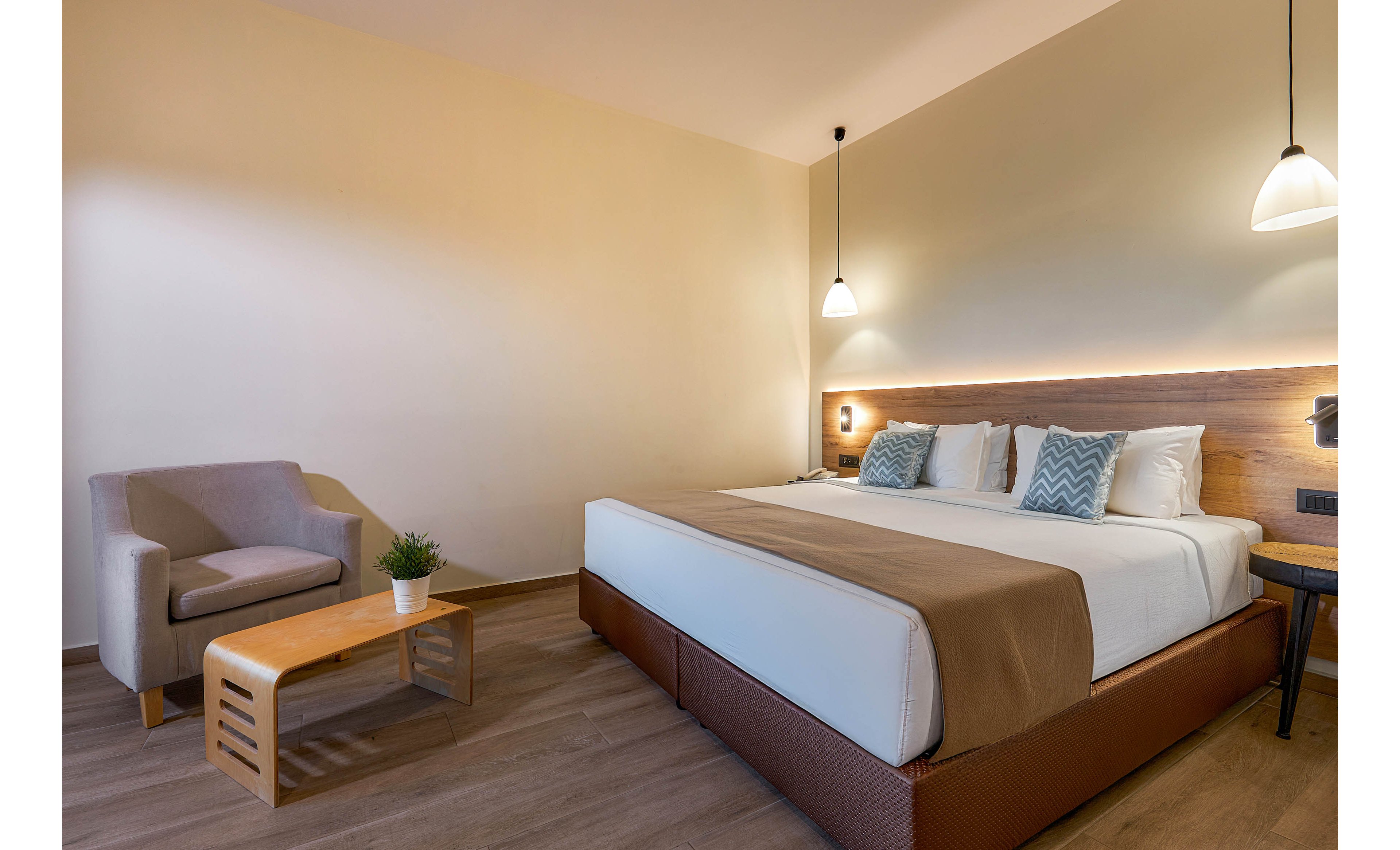 A spacious hotel room features a large king-sized bed with neutral bedding and decorative pillows. The setting includes a comfortable armchair, a wooden side table with a small potted plant, and warm pendant lighting. The room has a minimalist, contemporary style with soft beige and brown tones. No visible text or numbers are present in the image.