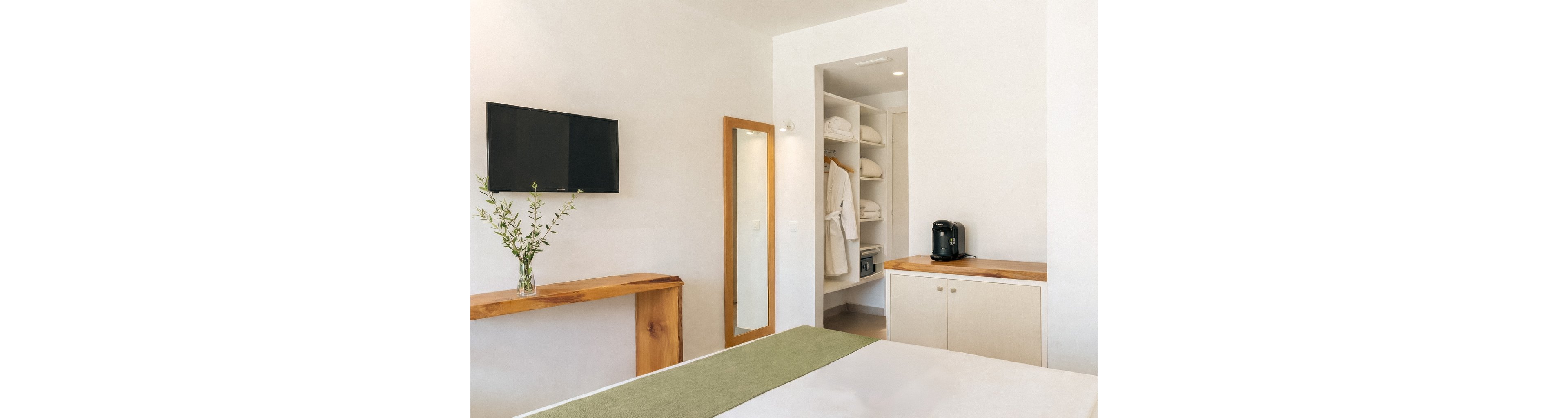 A contemporary hotel room features a neatly made bed with a green runner, a wall-mounted television, and a wooden console with a vase of greenery. The open closet displays white towels and bathrobes, while a coffee machine sits on a side cabinet. A tray with three colorful drinks and snacks is placed on the bed, creating a welcoming and relaxing atmosphere.