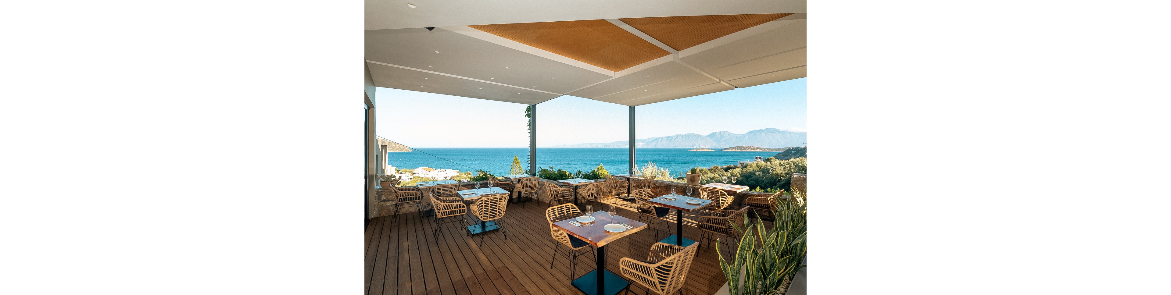 A stylish open air restaurant terrace features wicker chairs and wooden tables set for dining. The setting overlooks a stunning blue sea with distant mountains under a clear sky. The mood is relaxed and inviting, with natural light and a warm, earthy palette. No people are present, emphasizing the tranquil atmosphere. No visible text or numbers appear in the image.