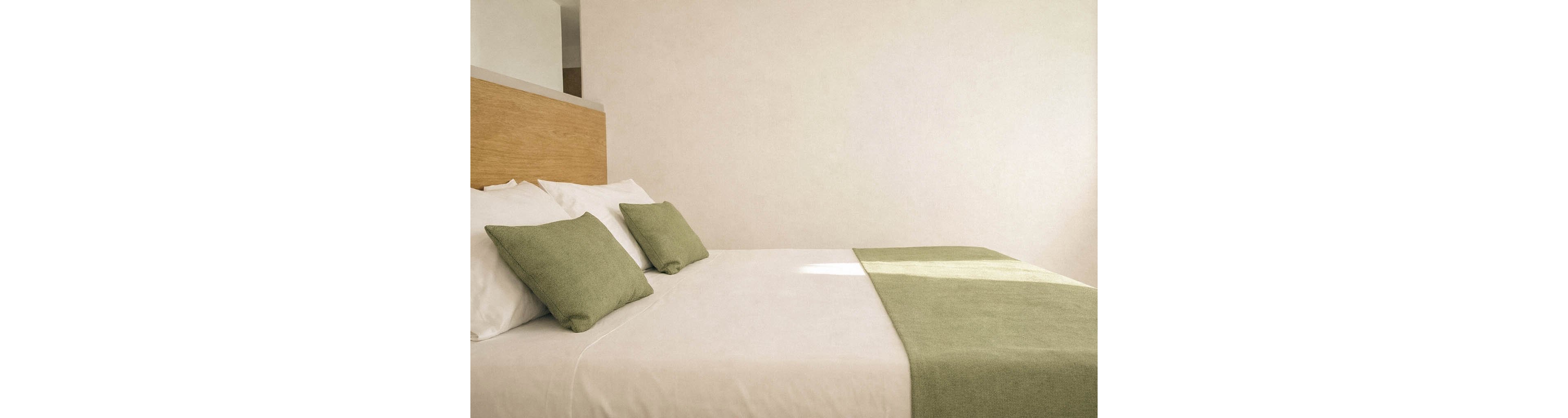 A neatly made bed with white linens and green pillows is the main focus of this minimalist hotel room. The setting is bright and airy, with sunlight casting soft shadows across the bed. The decor features a simple wooden headboard and a green runner, creating a calm and inviting atmosphere. No visible text or numbers are present in the image.