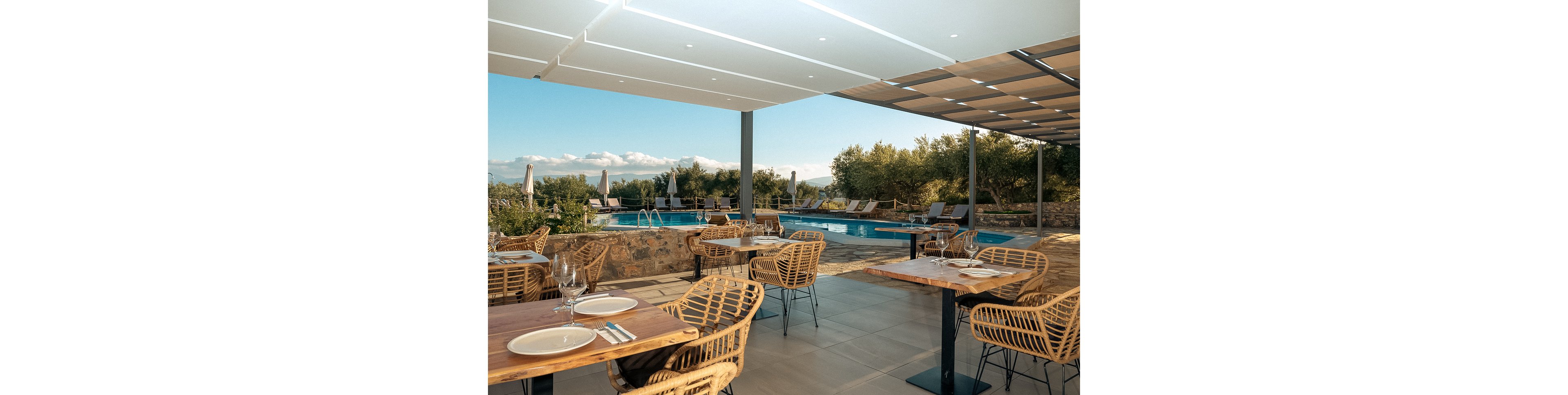 A stylish outdoor restaurant terrace features wooden tables and wicker chairs set for dining. The area overlooks a serene swimming pool surrounded by sun loungers and lush greenery, with mountains visible in the background. The setting is shaded by a modern pergola, creating a relaxed and inviting atmosphere. The overall mood is tranquil and sophisticated, ideal for leisure or vacation dining.