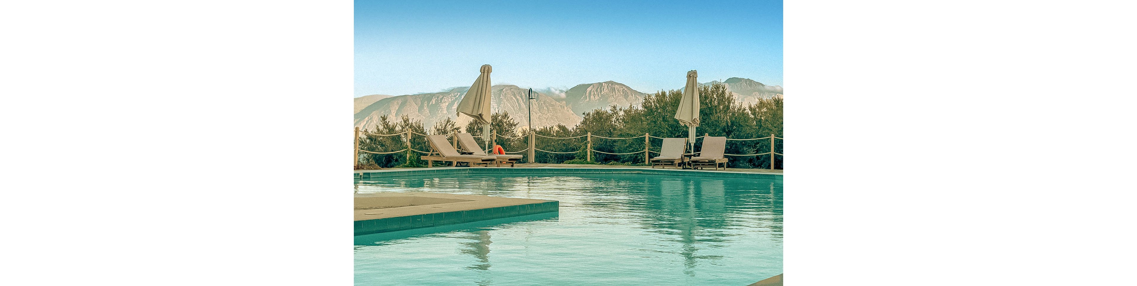 A tranquil outdoor swimming pool is featured in the foreground, surrounded by several empty lounge chairs and closed umbrellas. The setting is peaceful, with clear blue water reflecting the sky and distant mountains. The scene is bathed in soft morning light, creating a calm and inviting atmosphere. No people are present, emphasizing relaxation and solitude.